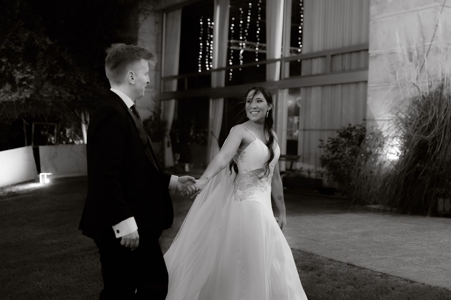 Black and white photo of a bride and groom holding hands outdoors at night during their wedding.