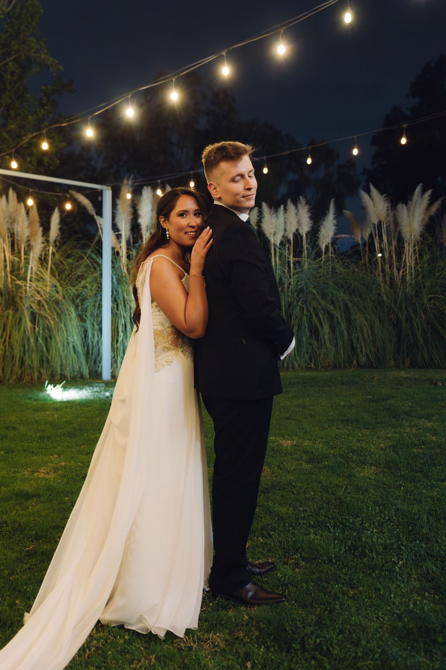 Bride and groom posing back-to-back under string lights with tall pampas grass at night wedding.