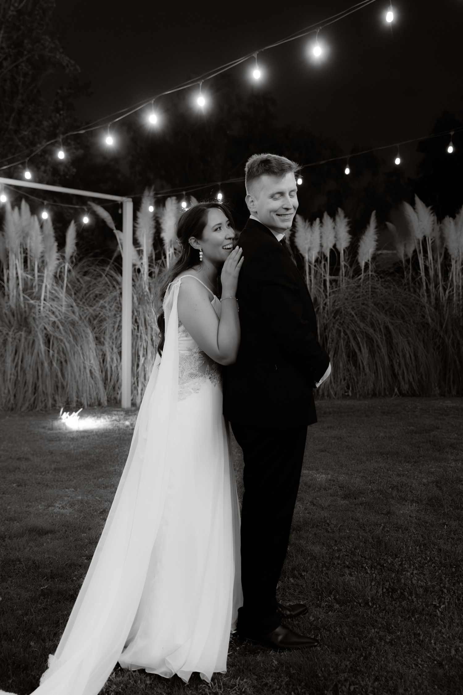 Black and white portrait of a bride and groom smiling under string lights surrounded by pampas grass.