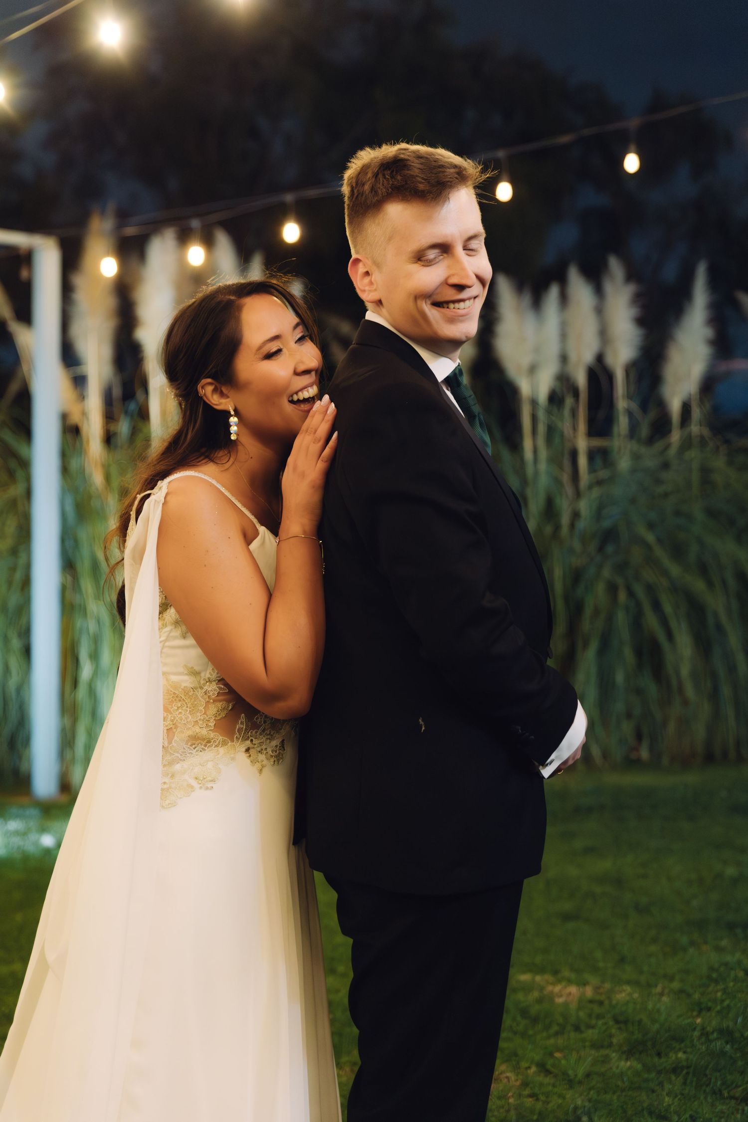 Bride laughing joyfully while standing behind smiling groom under string lights at outdoor wedding.