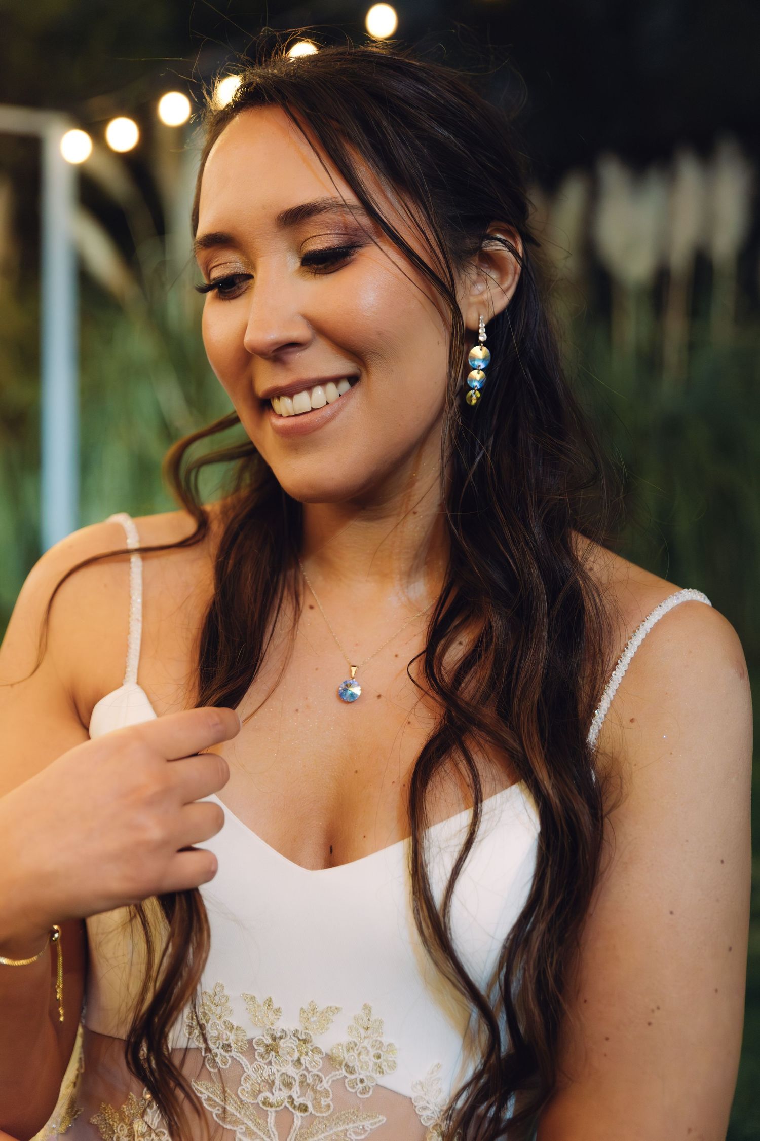 Beautiful bride with wavy dark hair smiling, wearing drop earrings and delicate necklace at wedding.