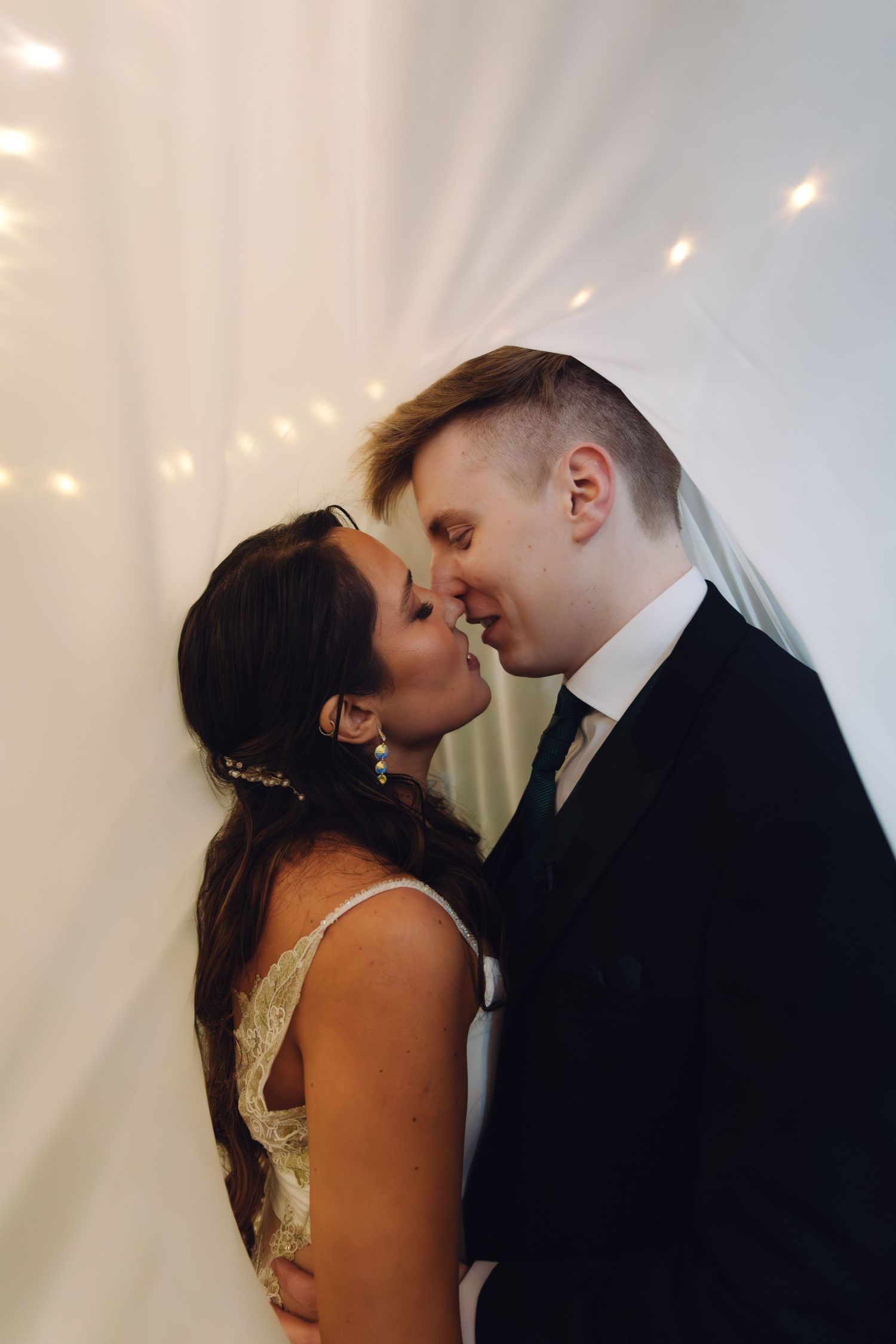 Bride and groom sharing a tender kiss against a white draped backdrop with soft fairy lights.