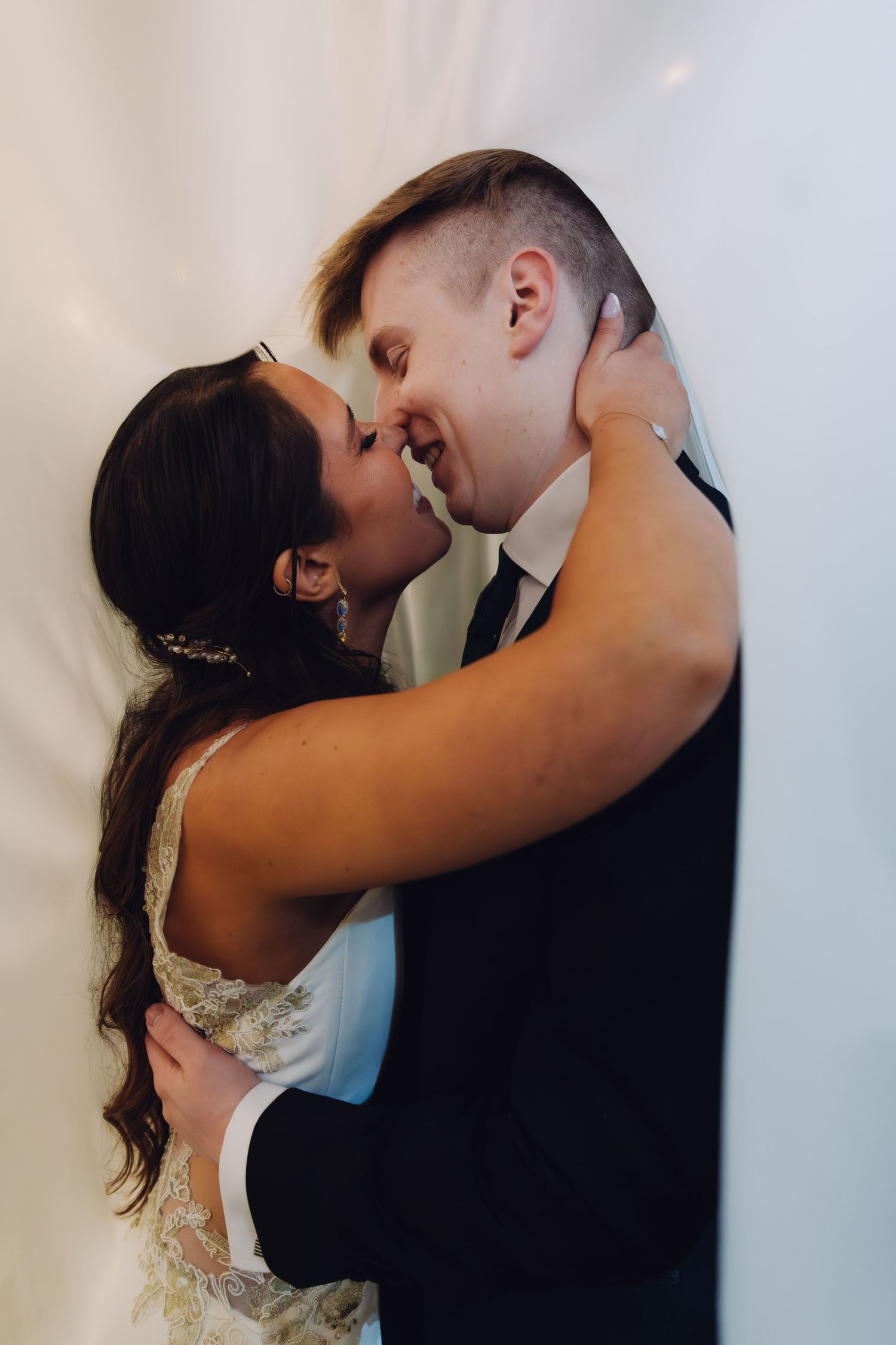 Bride and groom share a romantic almost-kiss, embracing tenderly against a white backdrop at their wedding.
