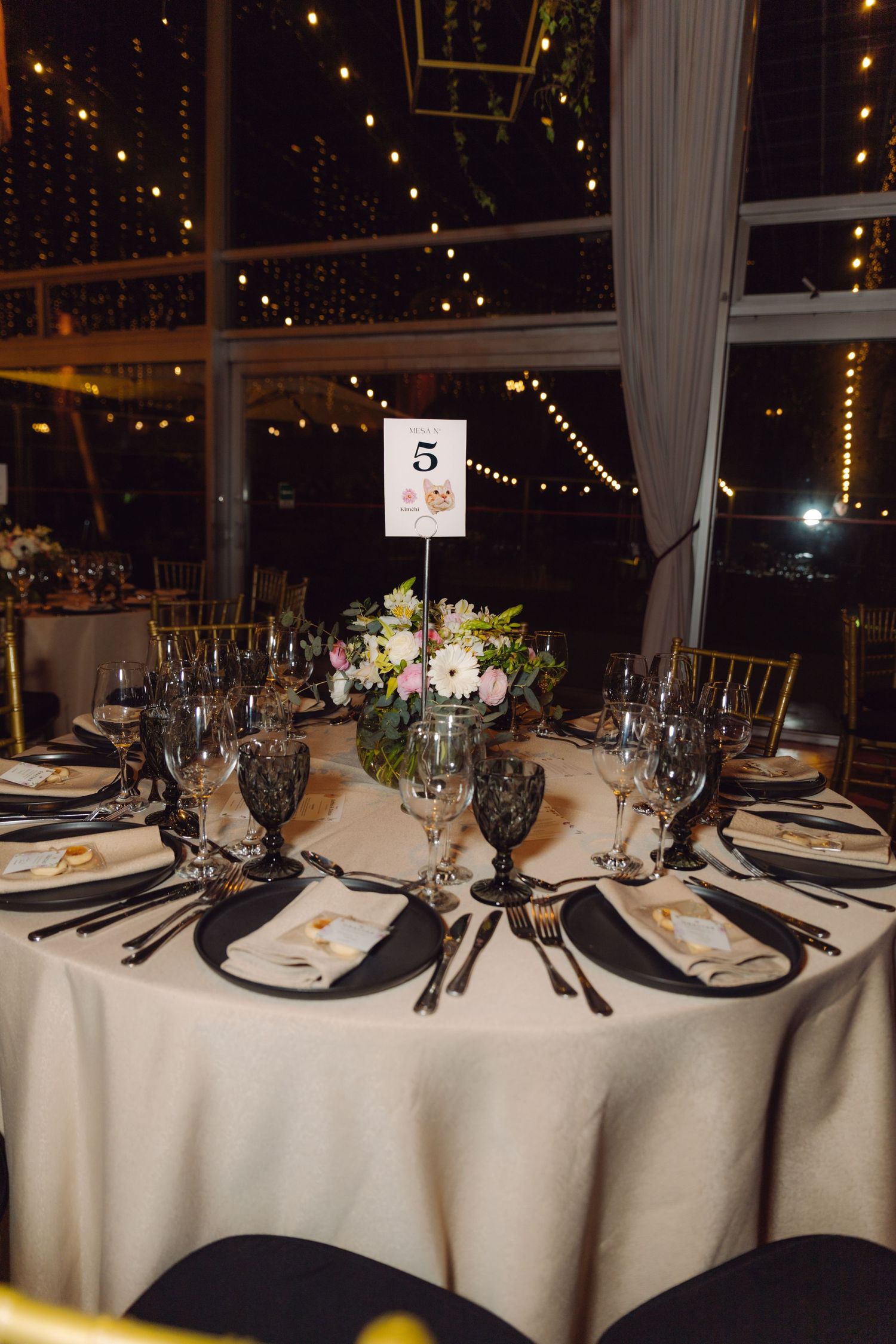 Elegant wedding reception table setting with floral centerpiece, number five card, and string lights in background.