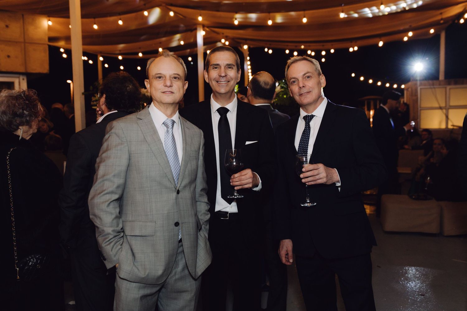 Three men in suits smiling at an evening wedding reception with string lights in the background.
