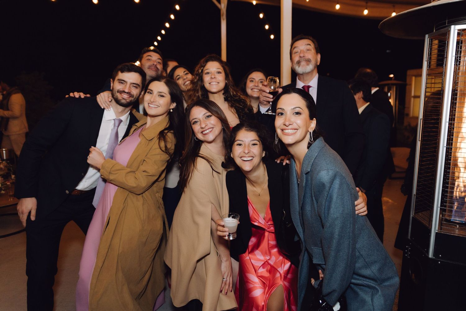 Group of wedding guests smiling together outdoors at a nighttime reception with patio heaters.