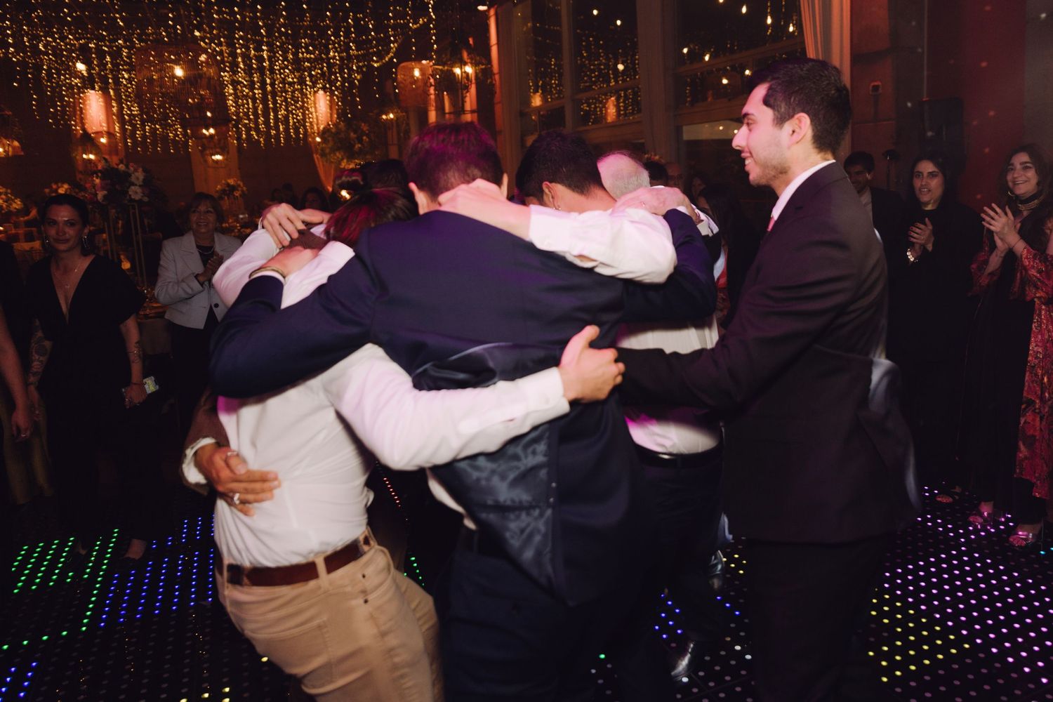 Group of male wedding guests hugging and celebrating together on a twinkling dance floor.