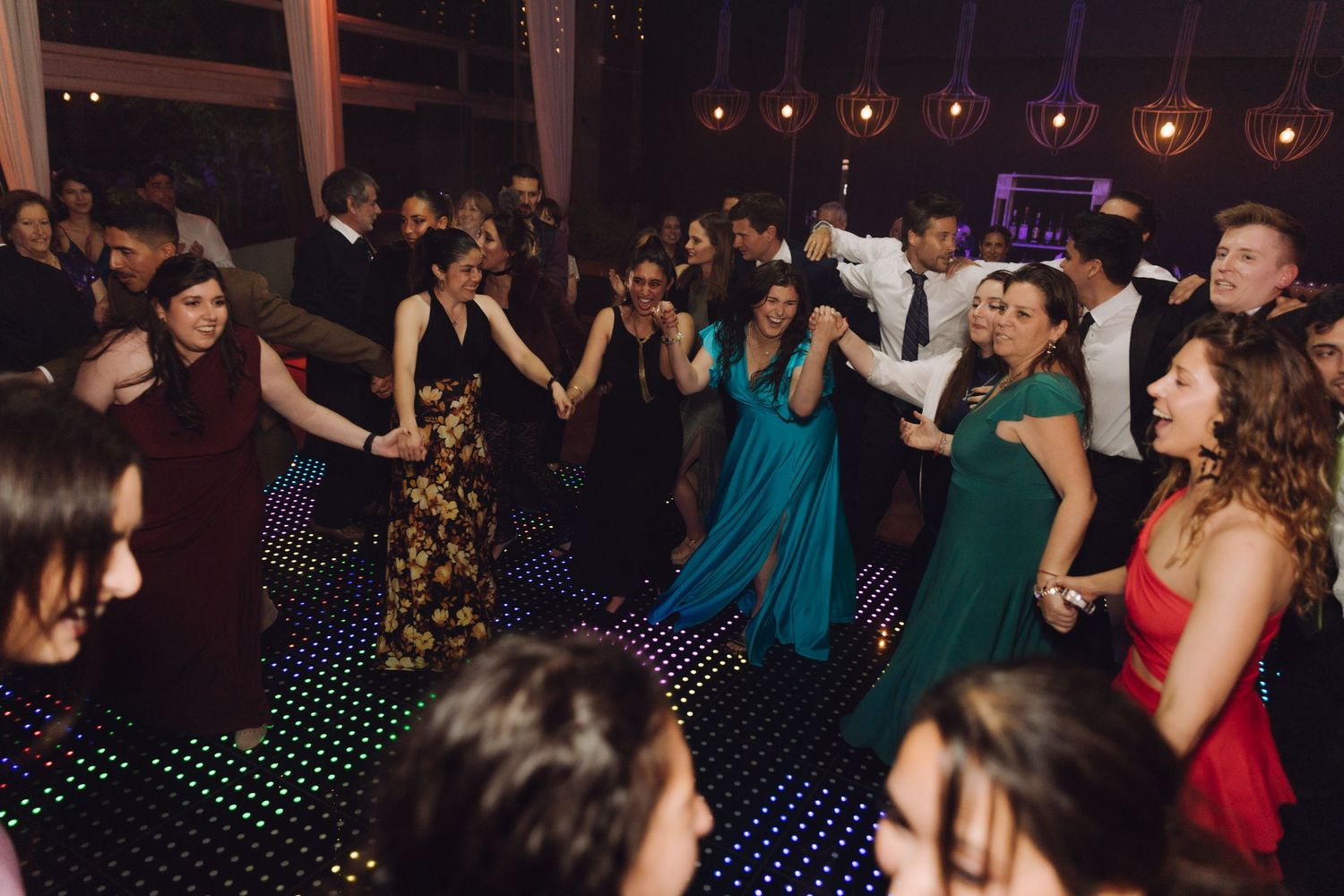 Wedding guests in formal attire form a chain dance on a twinkling LED dance floor.