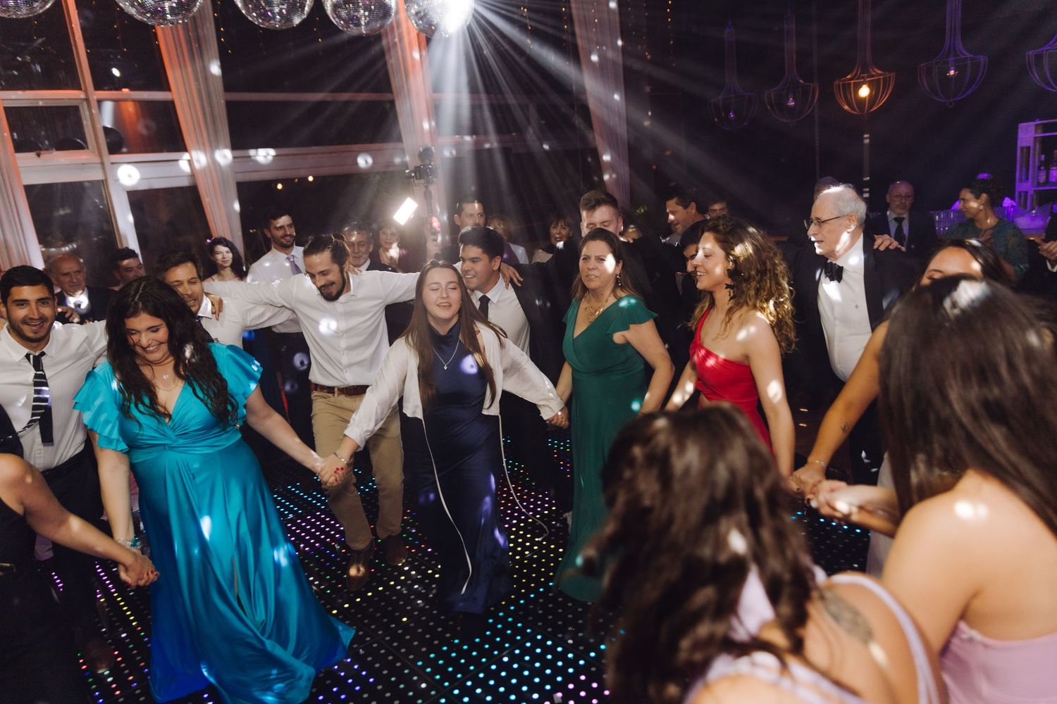 Wedding guests holding hands in a line dance on a black LED dance floor at a reception.