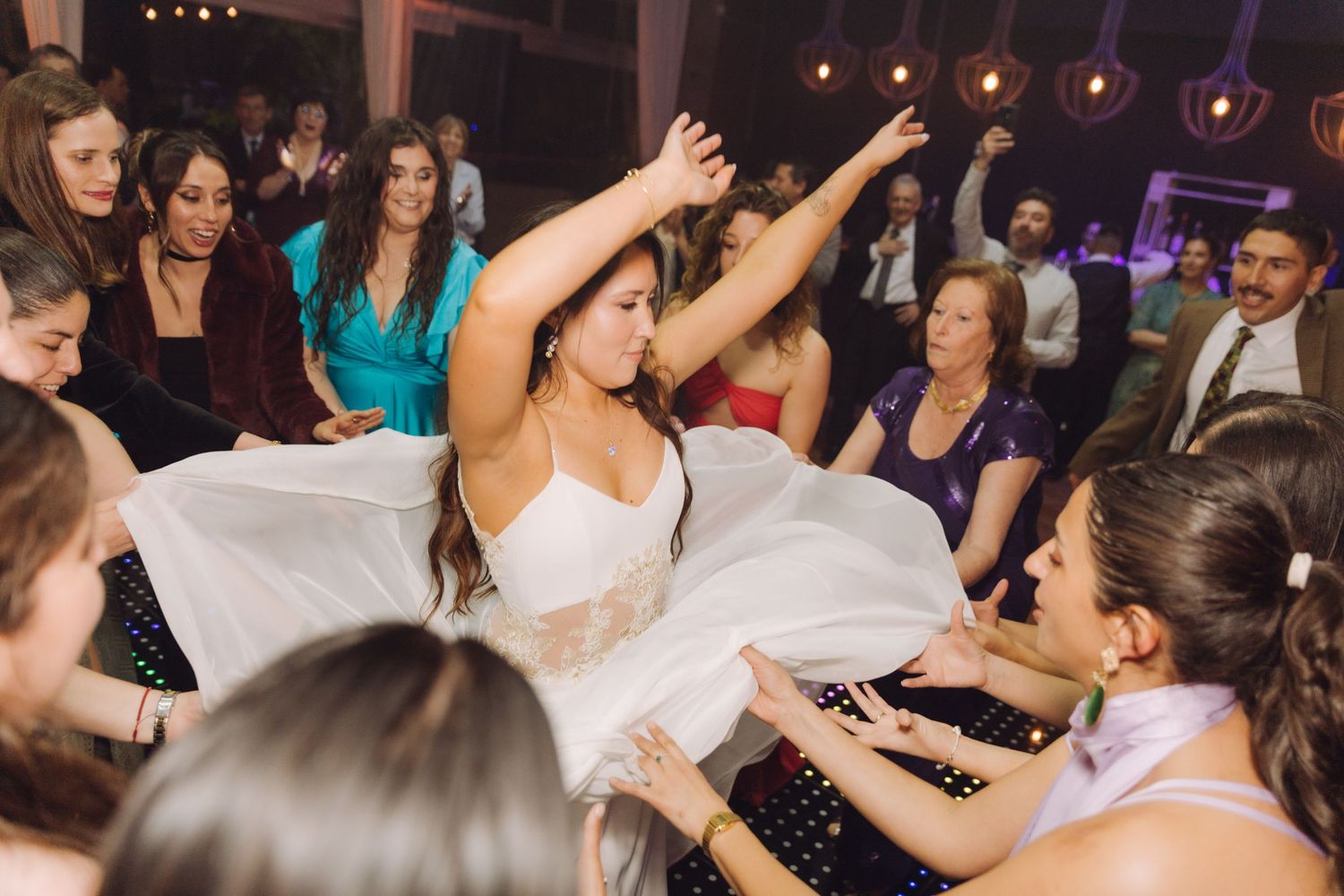 Bride dancing with arms raised while guests lift her wedding dress on the dance floor.