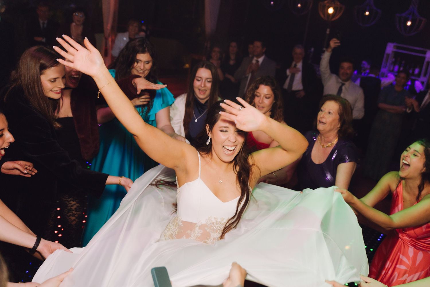 Joyful bride with arms raised being celebrated by guests on a wedding dance floor.