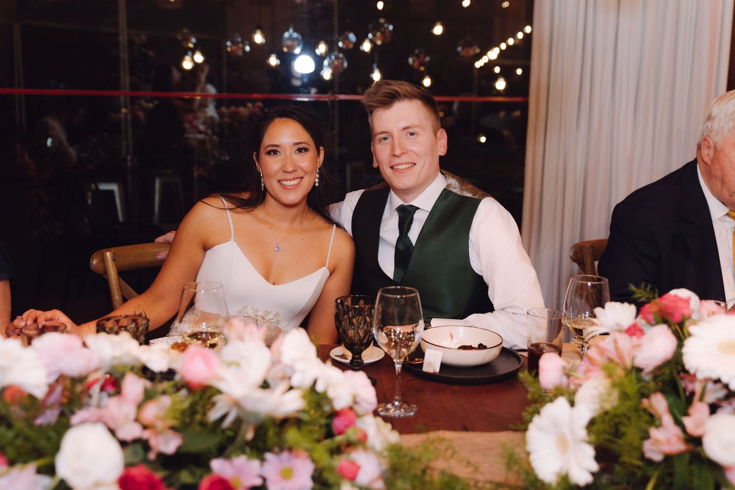Smiling bride and groom seated at a floral-decorated head table during wedding reception dinner.