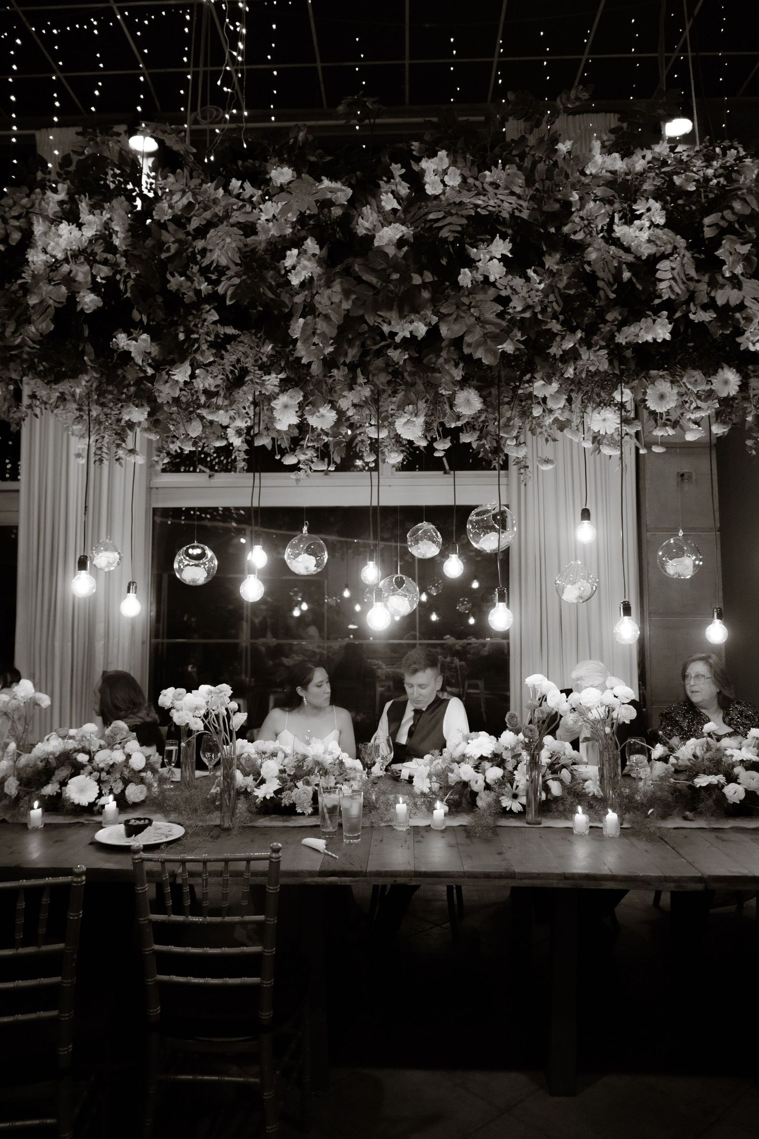 Black and white wedding reception with floral ceiling installation, hanging bulbs, and bride and groom seated at head table.