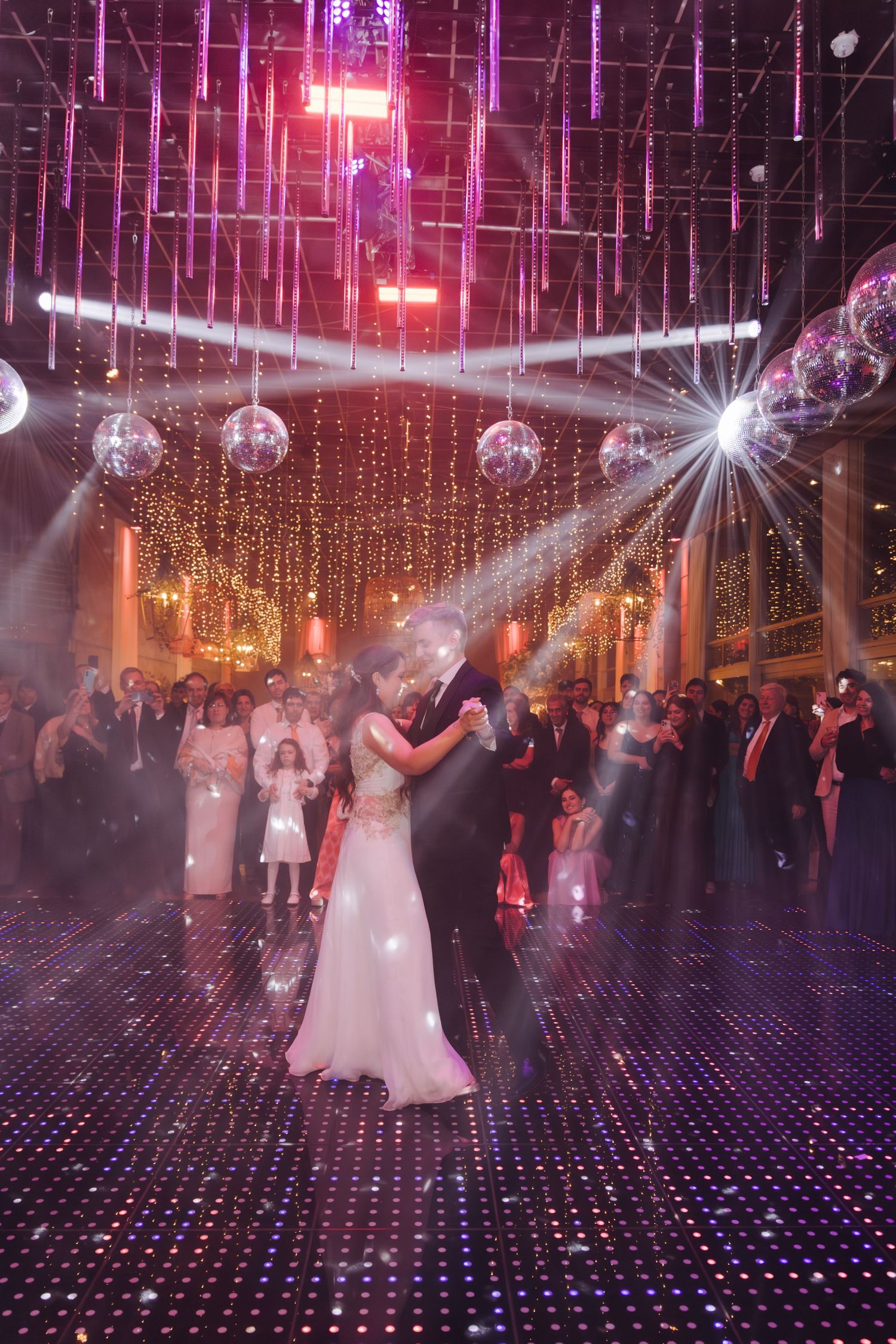 Bride and groom share first dance on sparkling dance floor under disco balls and colorful lights at wedding reception.