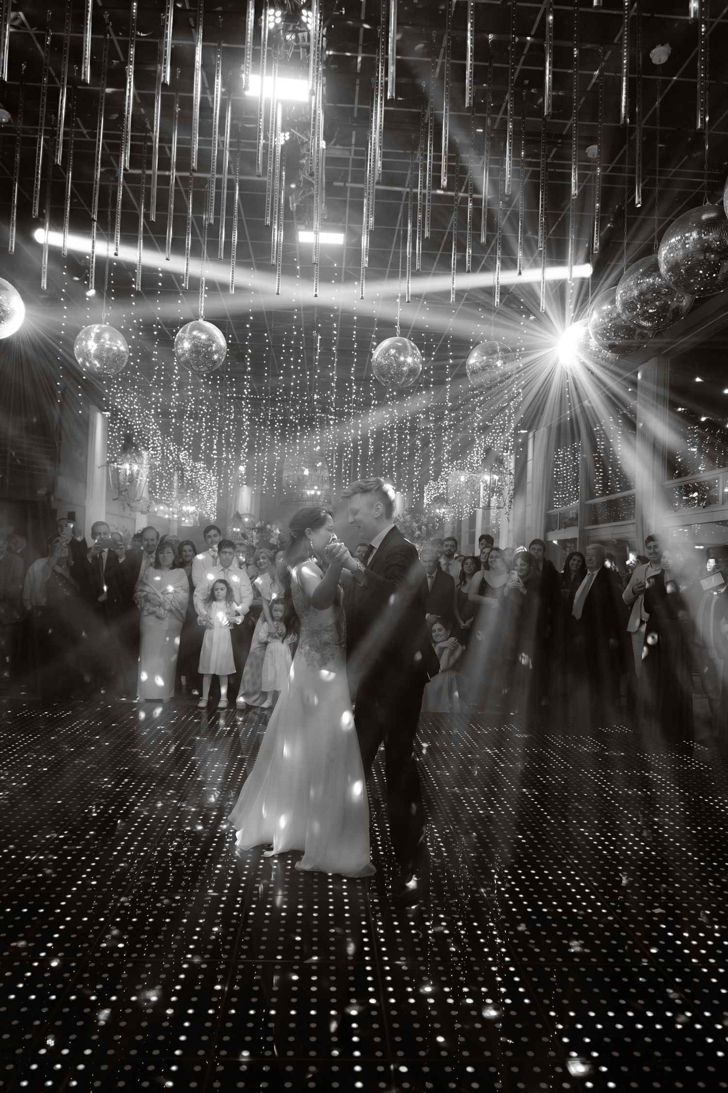 Black and white photo of newlyweds sharing first dance under glittering disco balls and light beams at wedding.