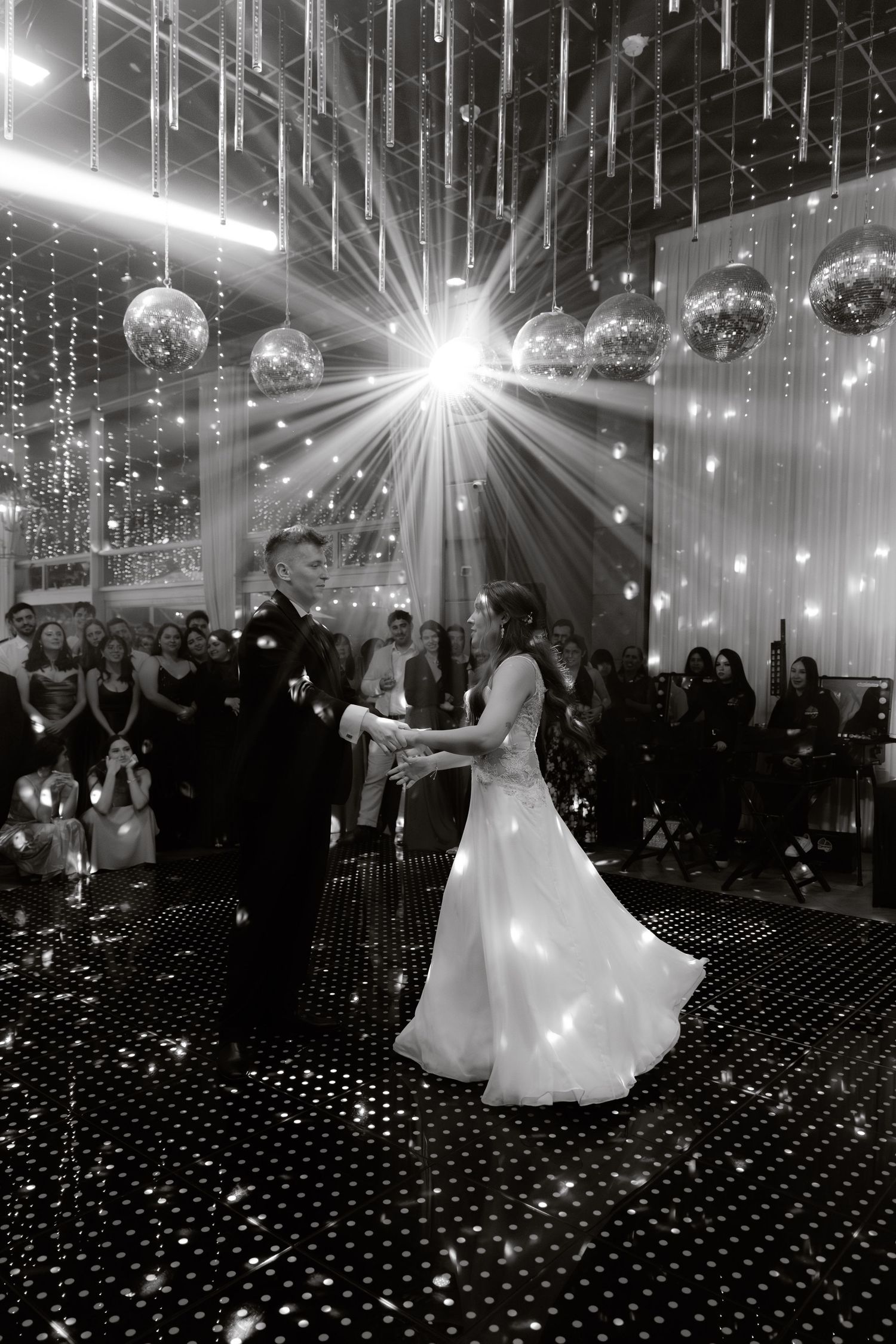 Black and white first dance photo of newlyweds on sparkling dance floor with disco balls and light rays.