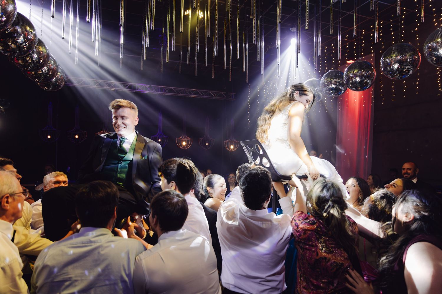 Bride and groom lifted by wedding guests in joyful hora dance celebration under colorful reception venue lights.