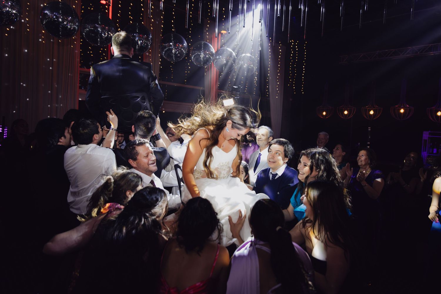 Bride and groom kissing while being lifted by cheering wedding guests during lively reception party celebration.