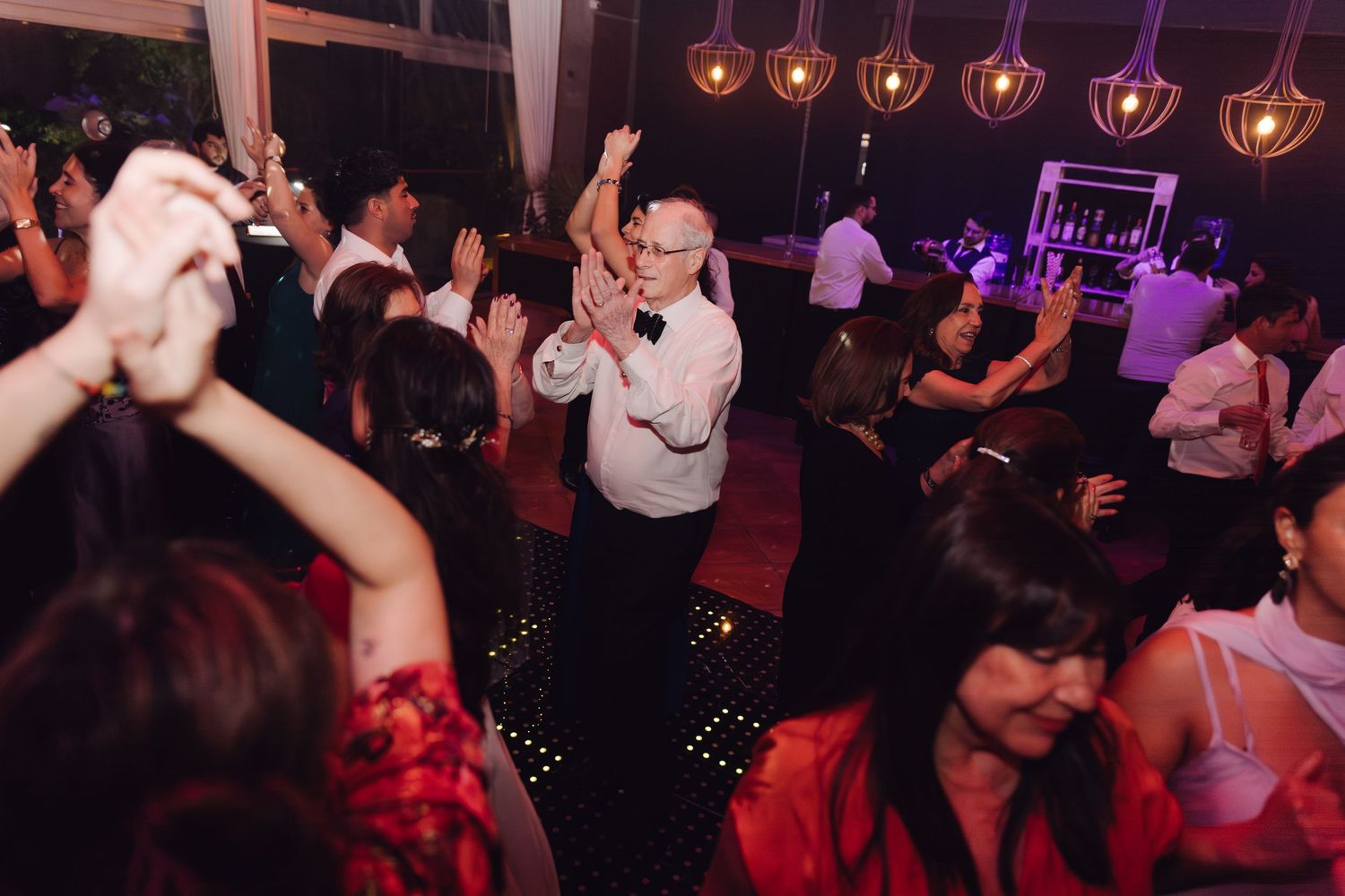 Wedding guests dancing energetically on star-lit dance floor with older gentleman raising hand in celebration.