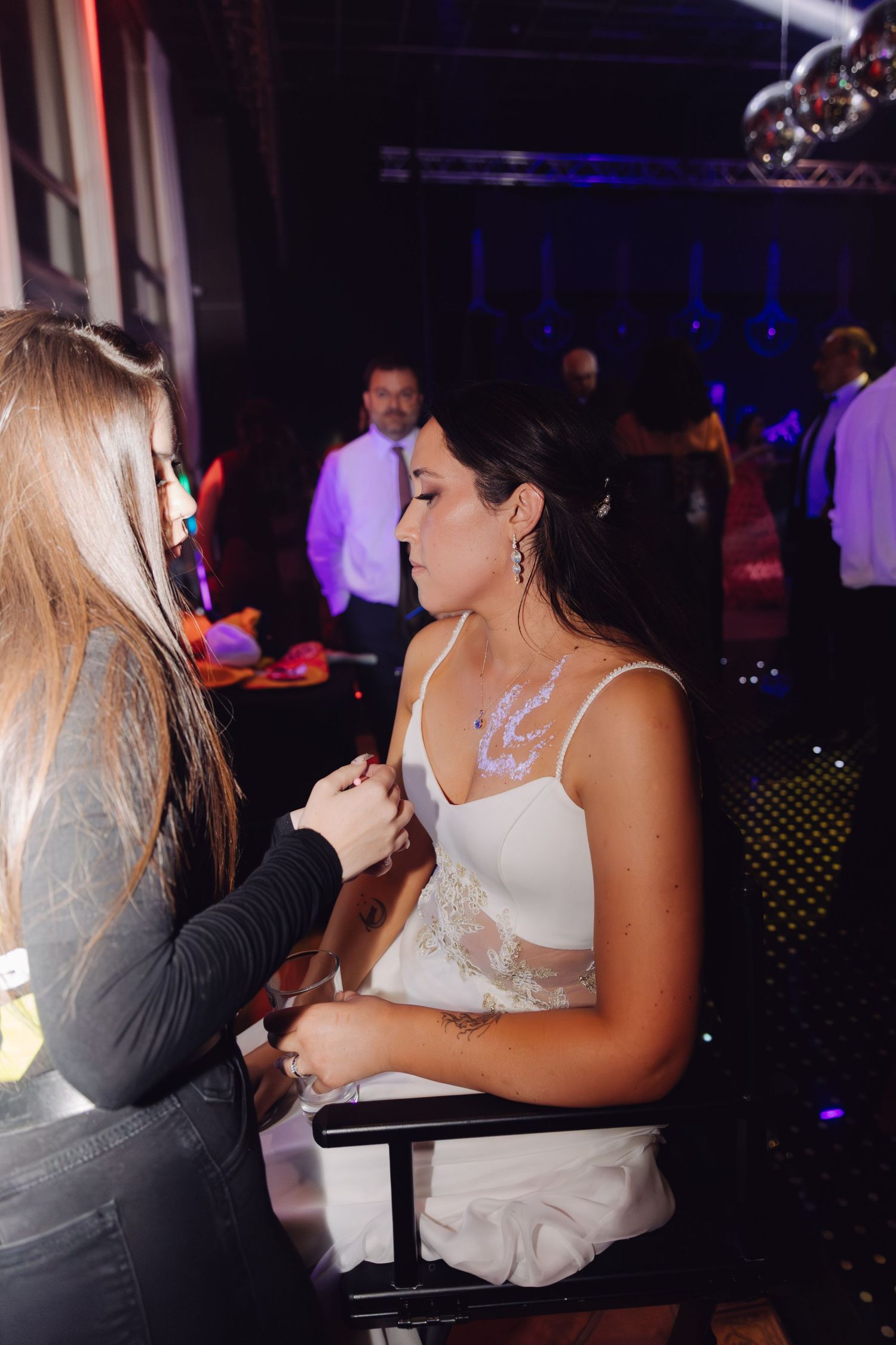 Makeup artist applying glitter body art to a bride in a white dress at a wedding reception.
