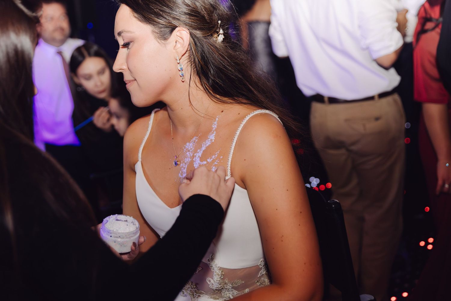 Bride in white dress with purple glitter body art holding a cup at a wedding party.