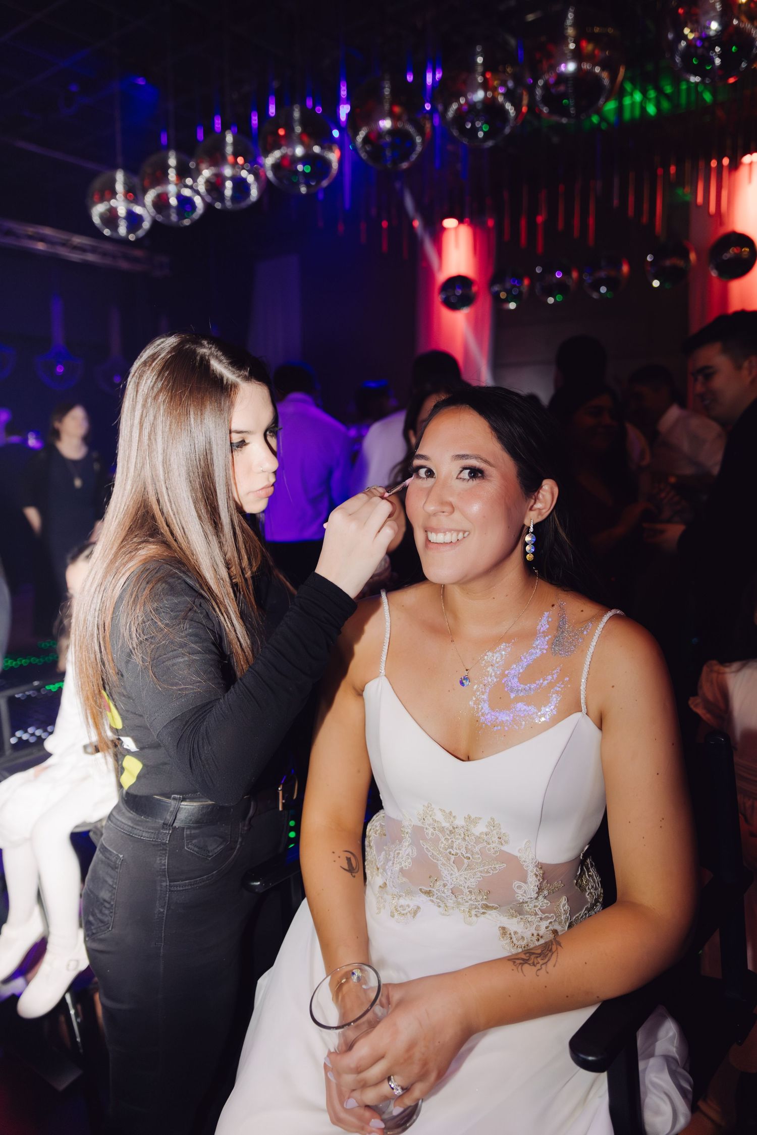 Makeup artist touching up bride's makeup on wedding reception dance floor with colorful lights.