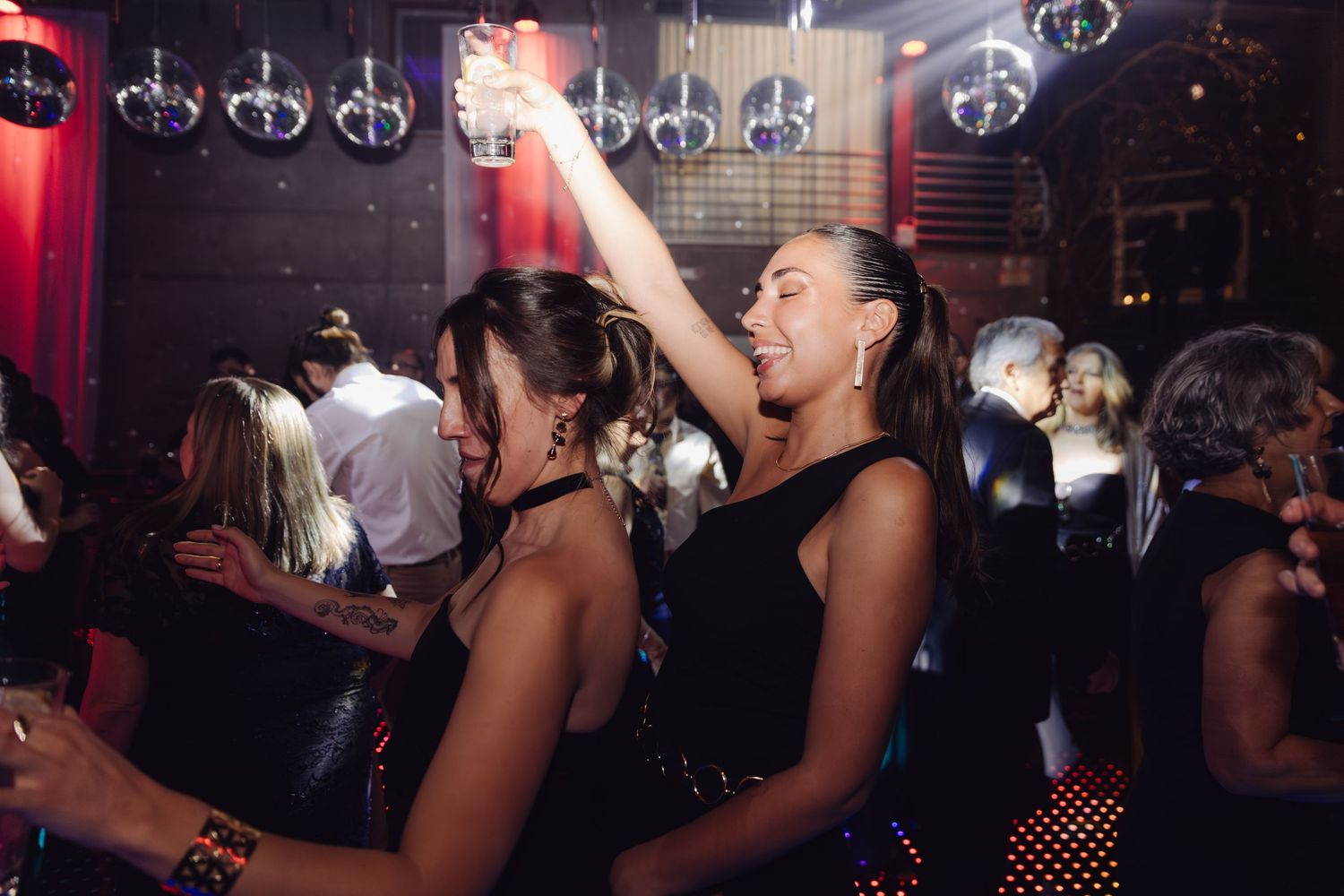 Two women in black dresses celebrating at lively wedding reception party with raised drinks.