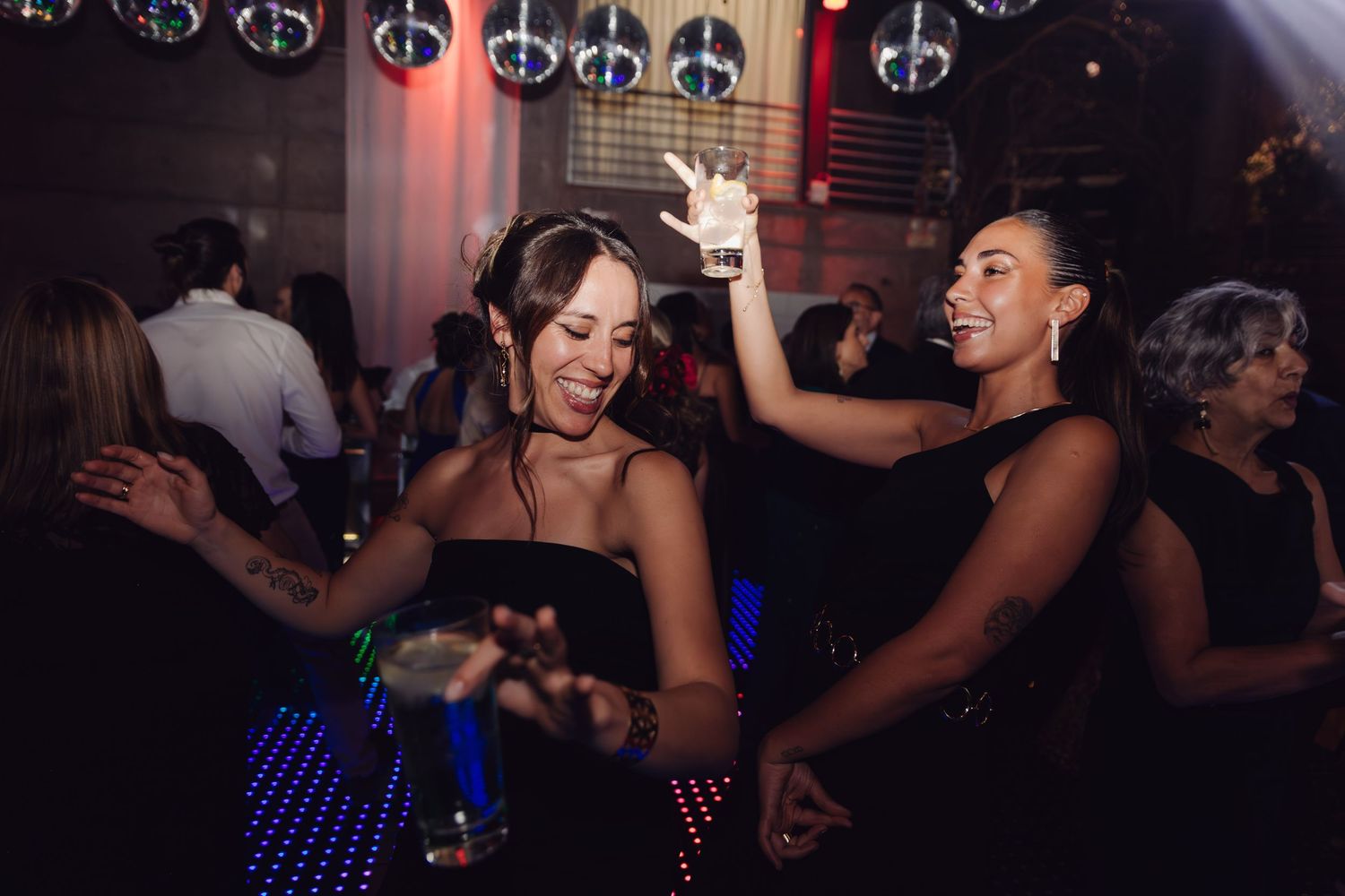 Two women in black dresses dancing joyfully at wedding reception with disco ball lighting.