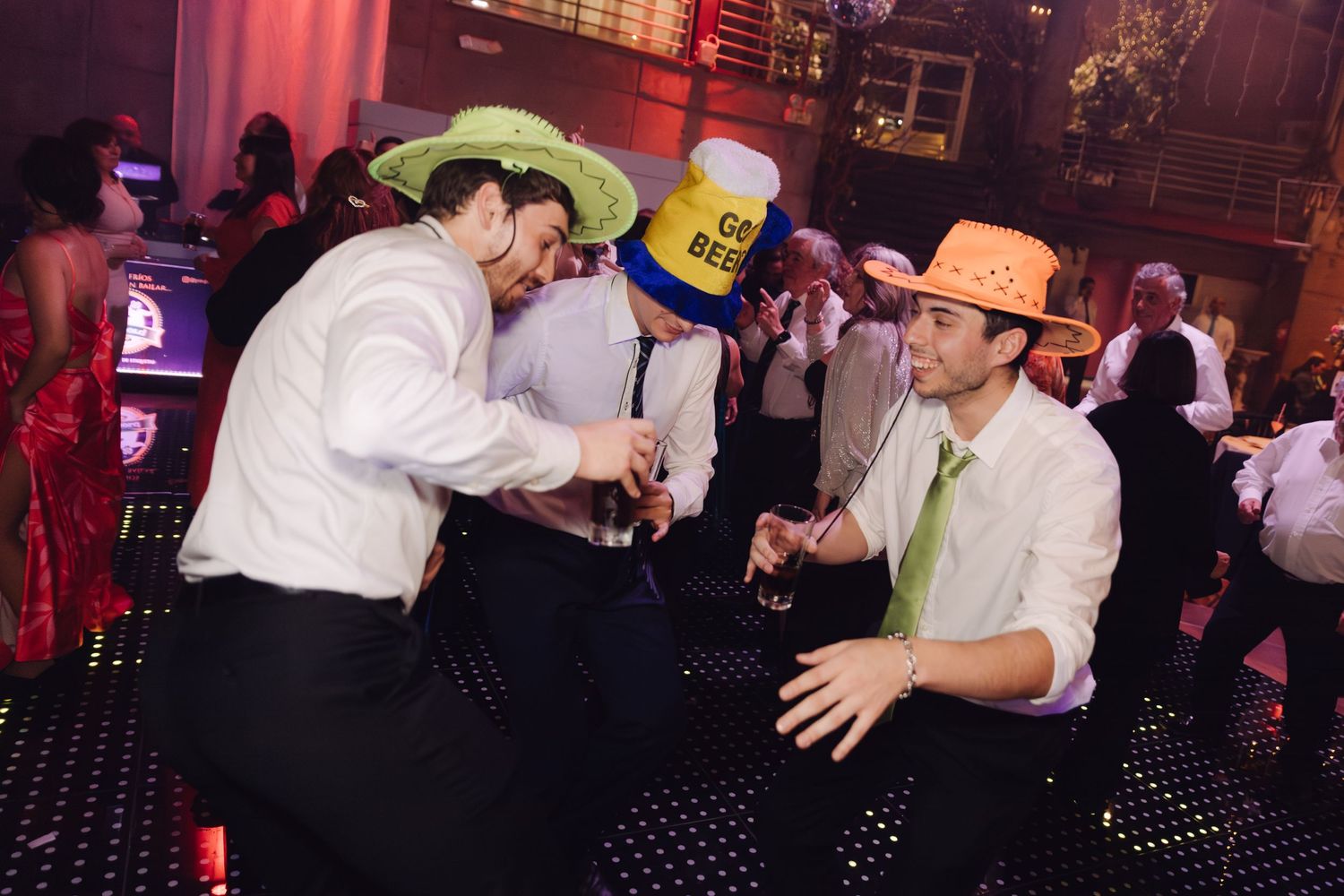 Groomsmen wearing novelty hats dancing playfully on illuminated dance floor at wedding reception.