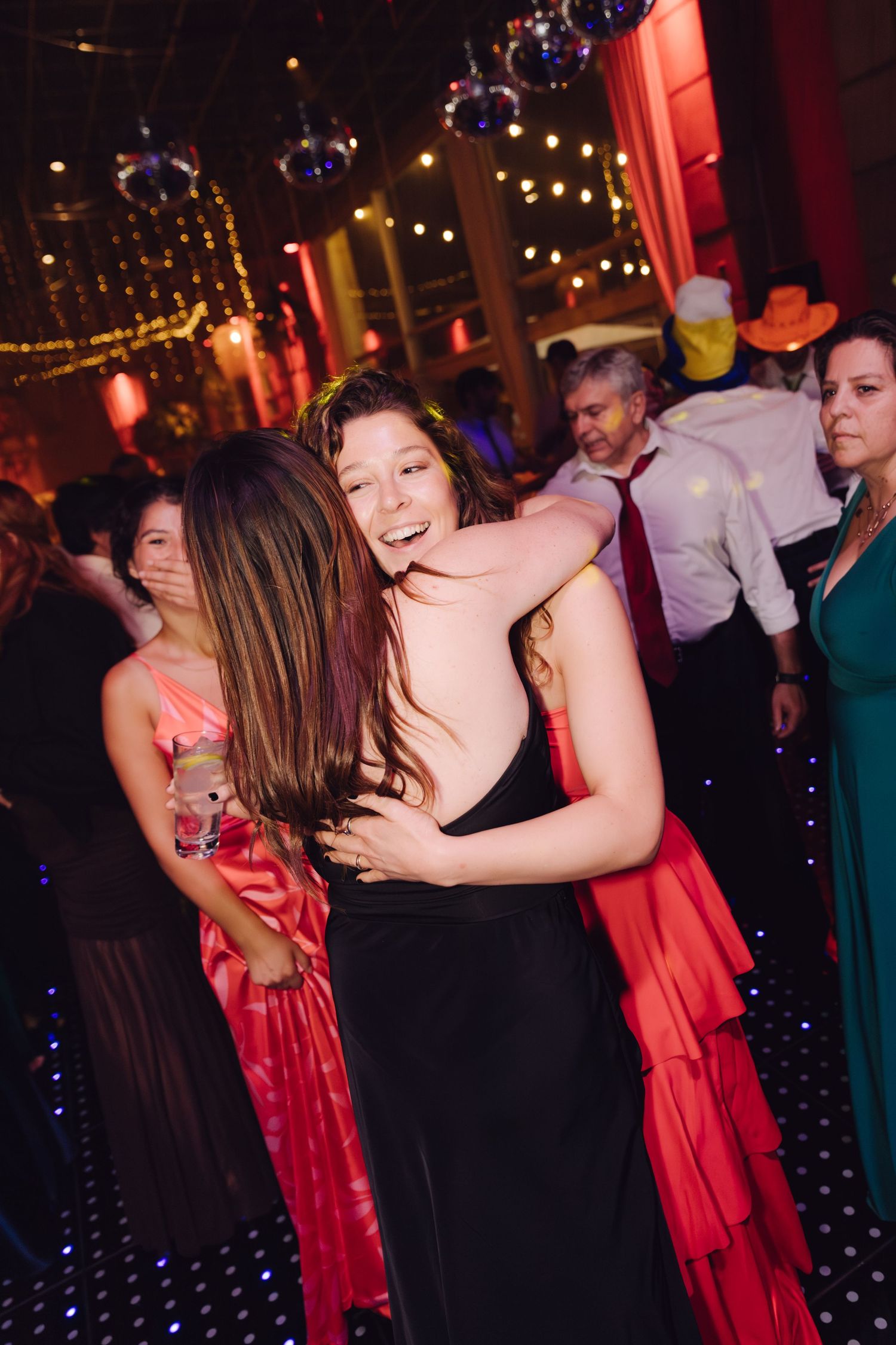 Two young women in elegant dresses share a joyful hug on a decorated dance floor at a formal event.
