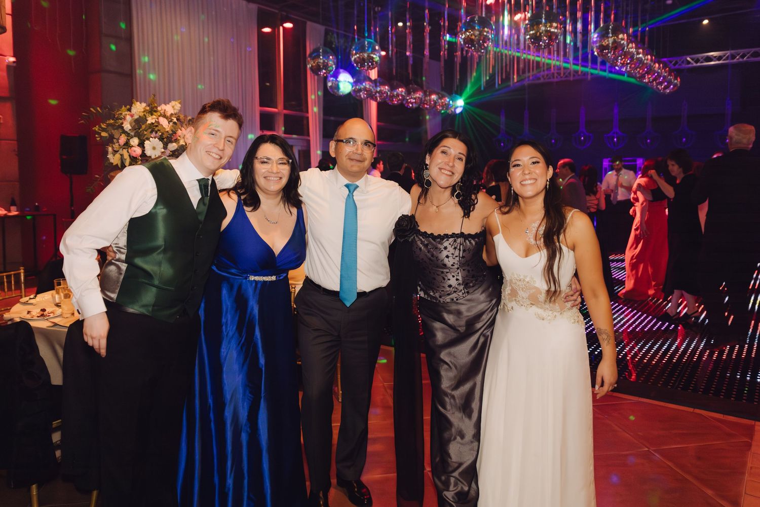 Group of five smiling guests posing together at an elegant wedding reception with colorful lighting.