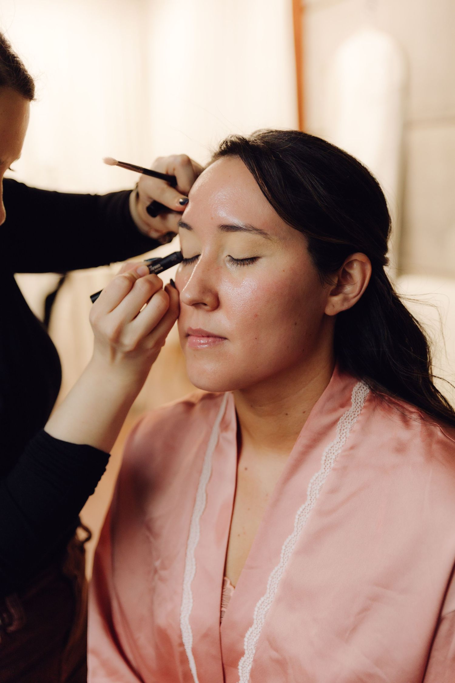 Makeup artist applying eyeshadow to bride wearing pink satin robe during wedding preparation.