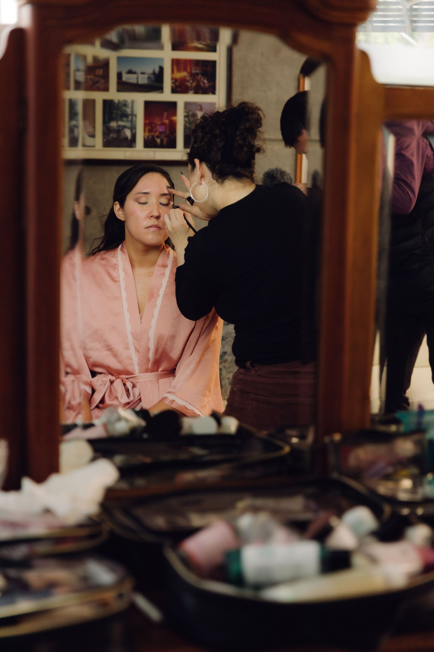 Mirror reflection of bride in pink robe getting makeup applied by artist at vanity table.