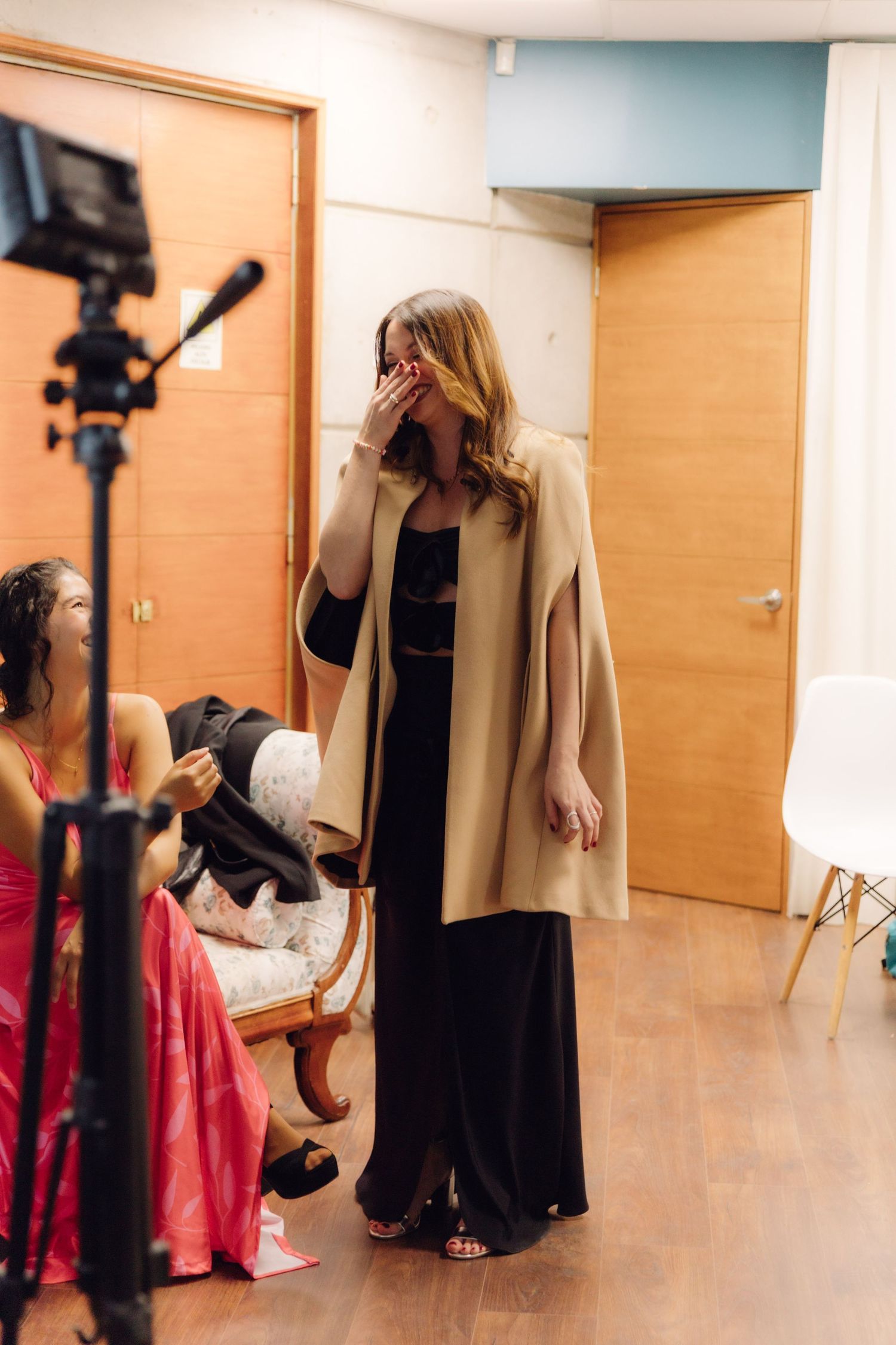 Emotional wedding guest in camel cape covering mouth while watching bridal preparations indoors.