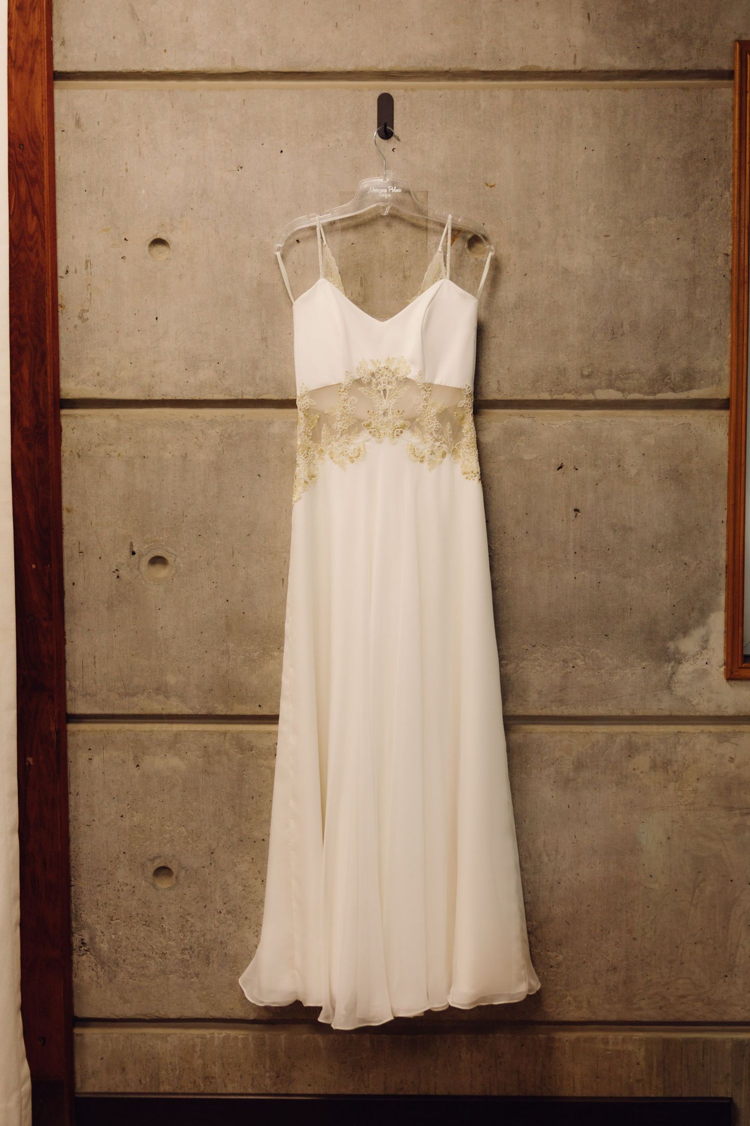 Elegant white wedding gown with gold lace applique detail hanging on concrete wall.