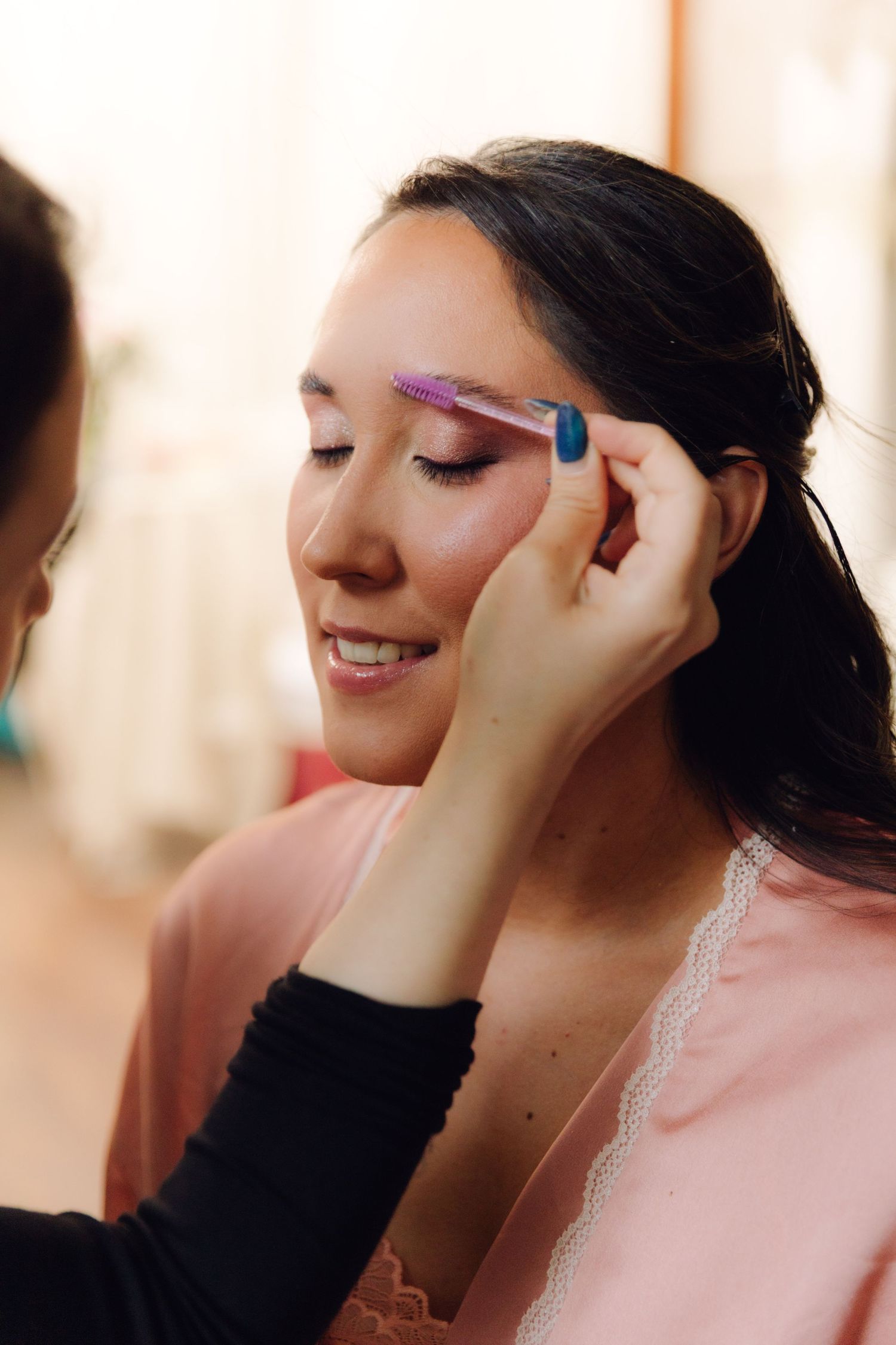 Smiling bride in pink robe having eyebrows groomed by makeup artist before wedding ceremony.