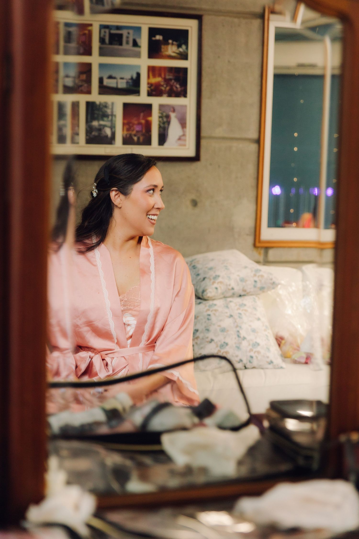 Happy bride in pink satin robe smiling at mirror reflection during wedding morning preparation.