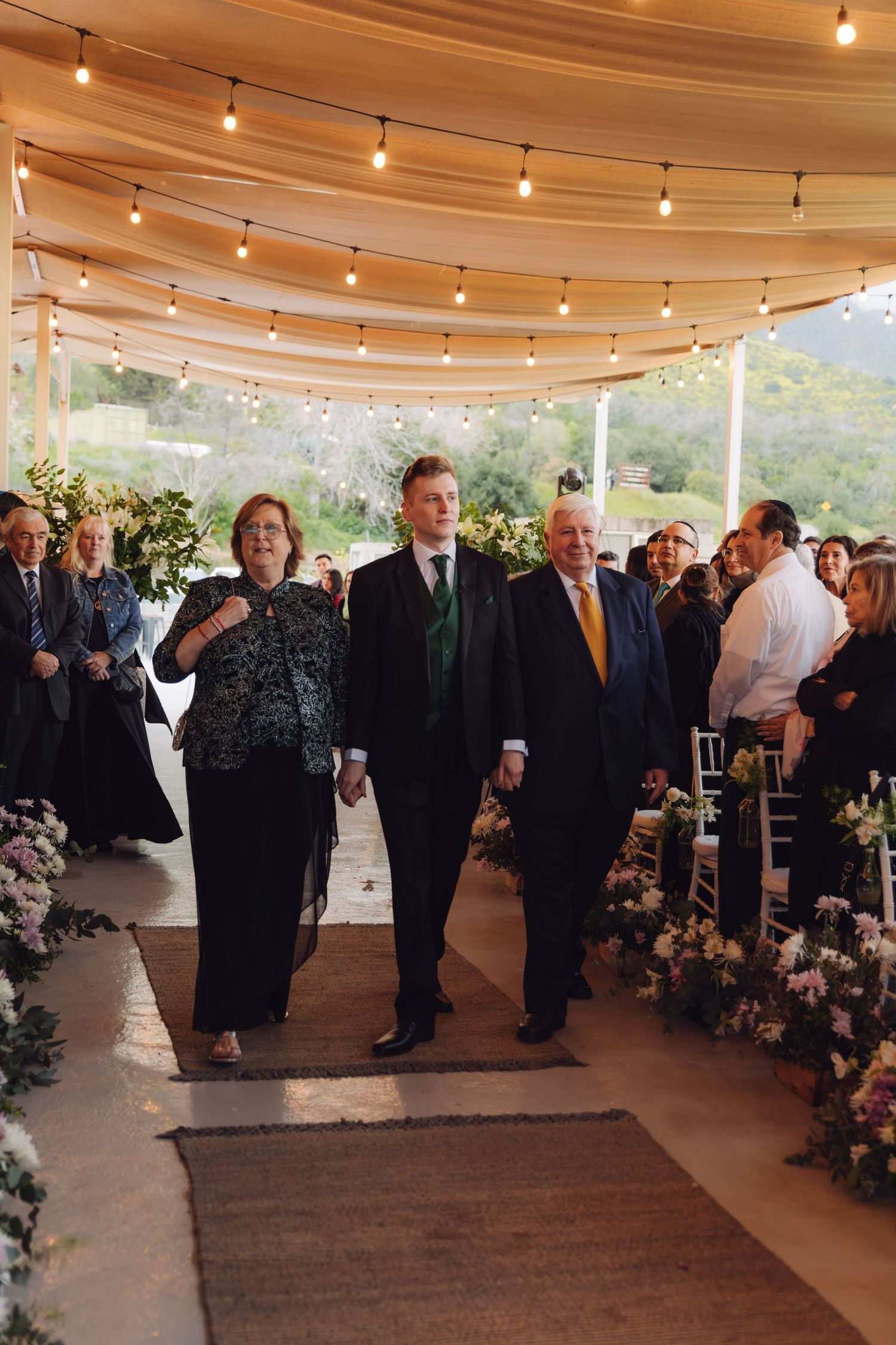 Groom walks down wedding aisle escorted by parents under string lights.