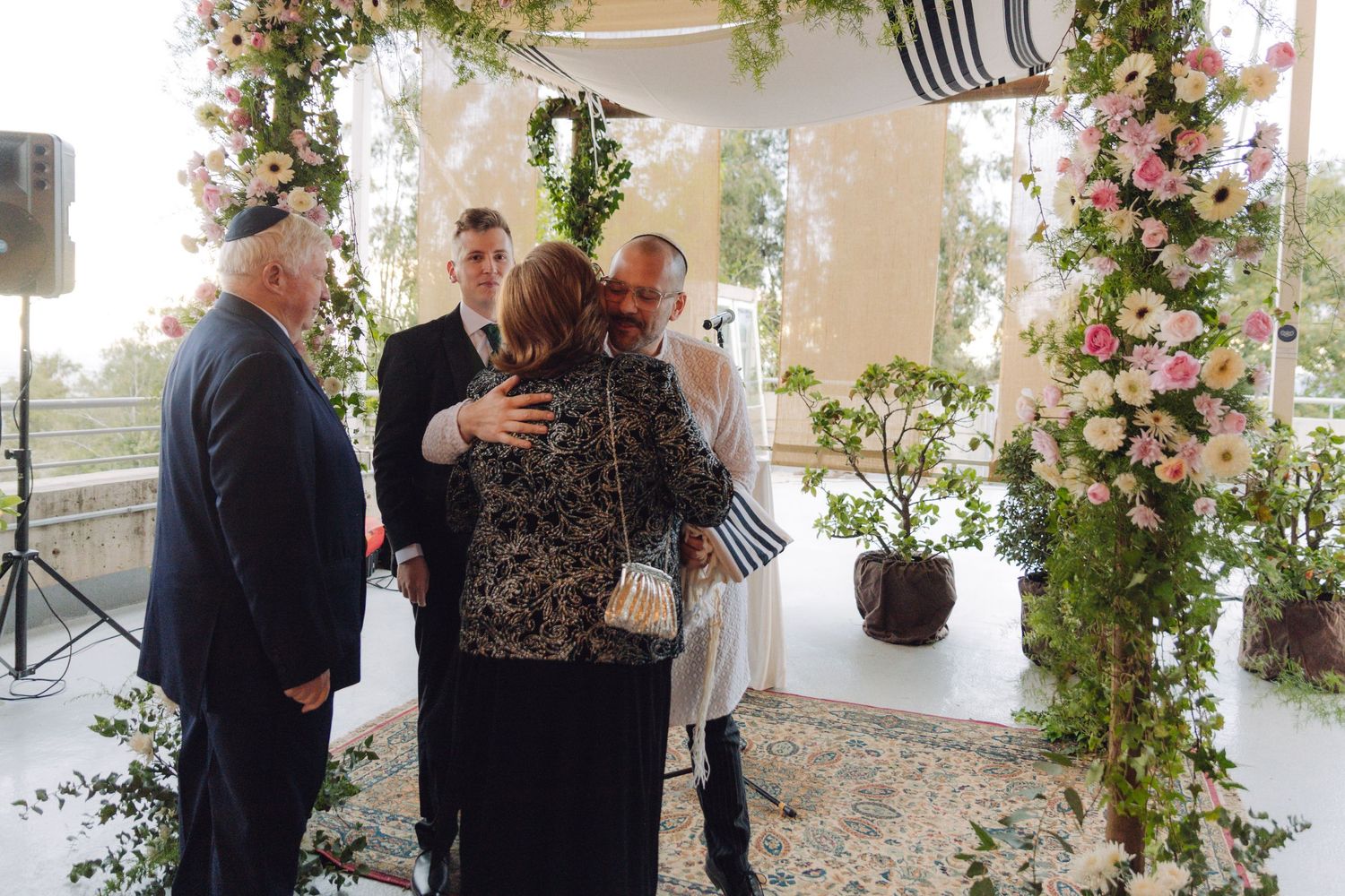 Family members share warm embraces at floral chuppah after Jewish wedding ceremony.