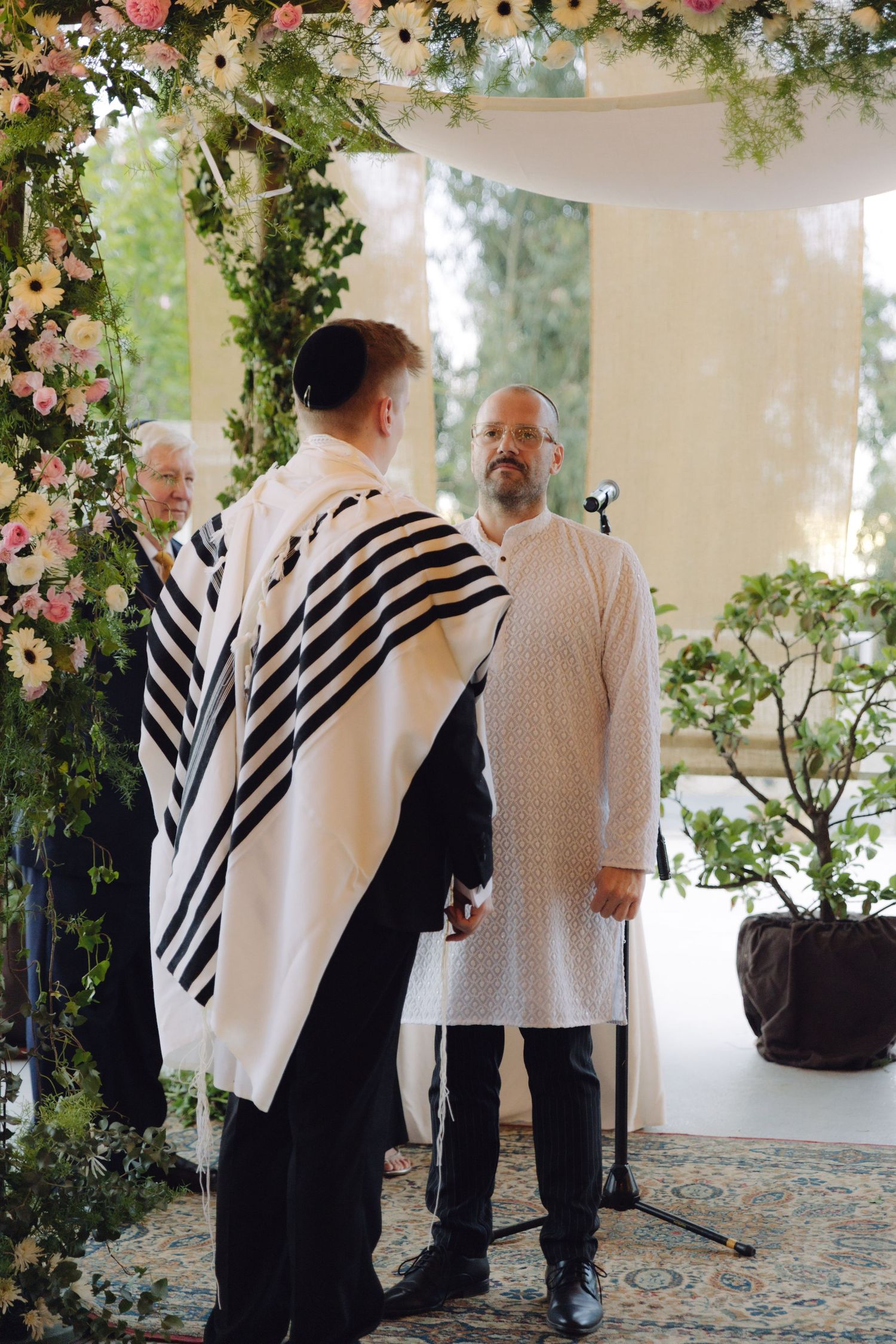 Groom wearing tallit stands at floral chuppah during Jewish wedding ceremony.