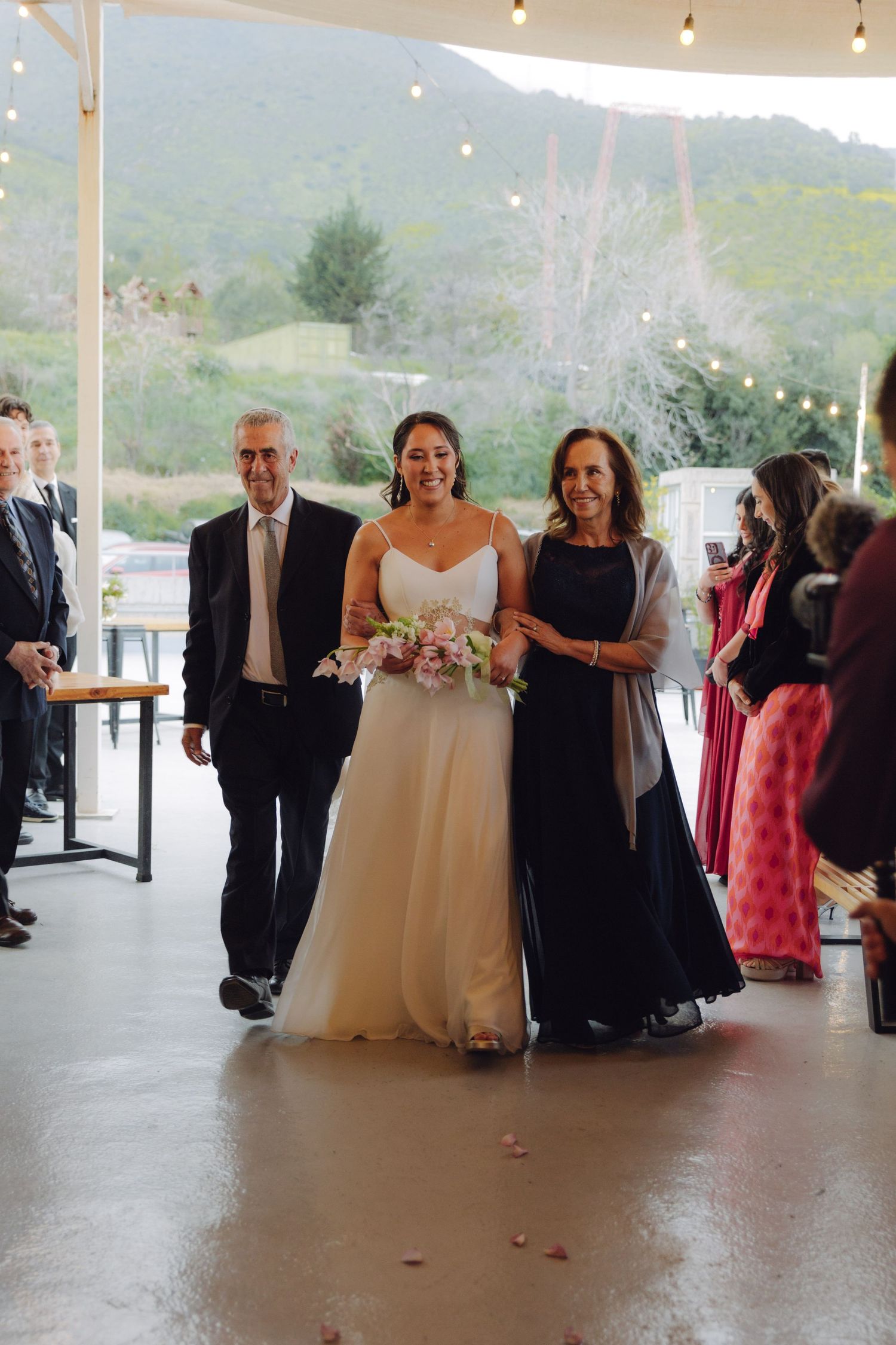 Smiling bride walks down outdoor wedding aisle escorted by both parents.