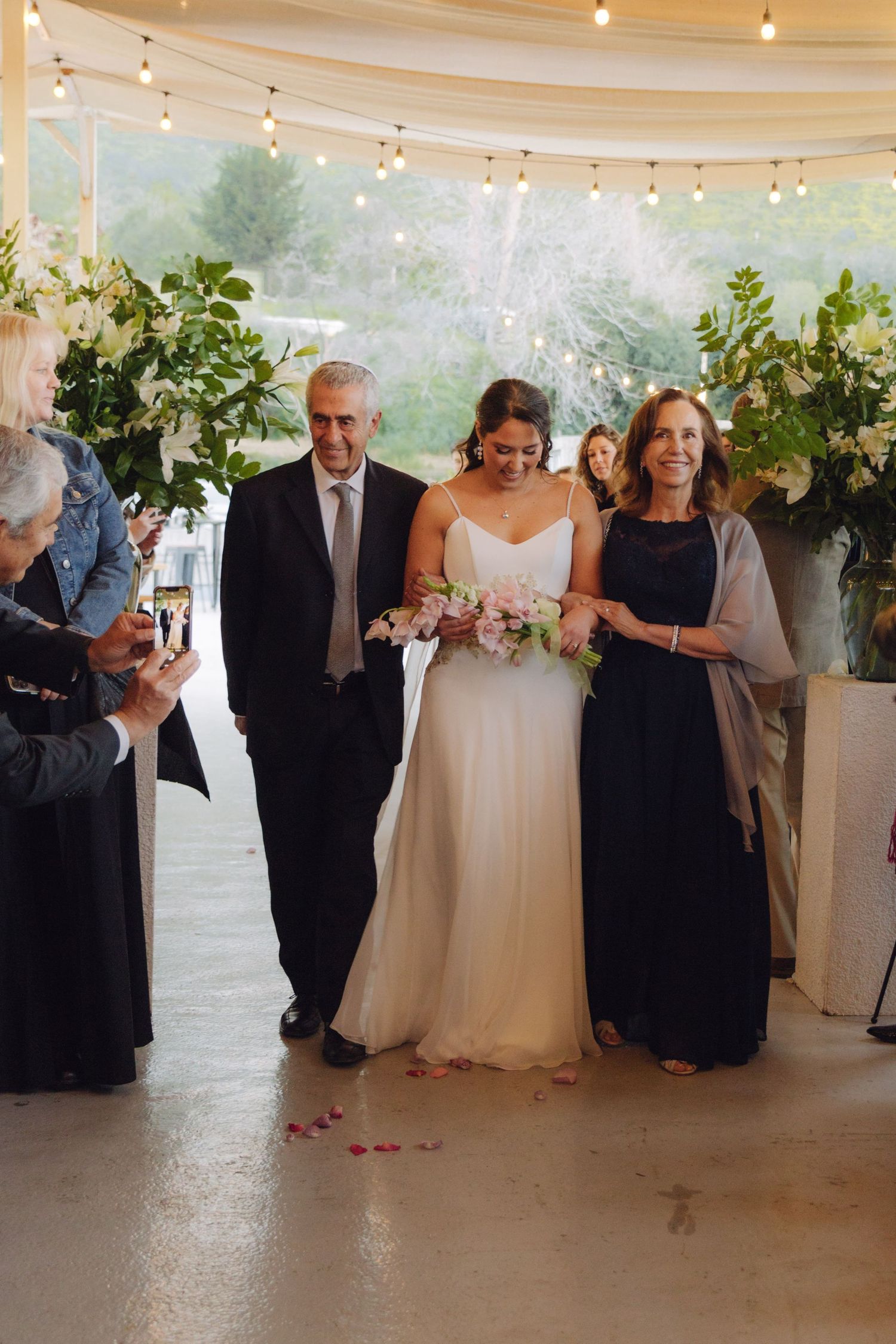 Bride in white gown walks down aisle with parents holding pink bouquet.
