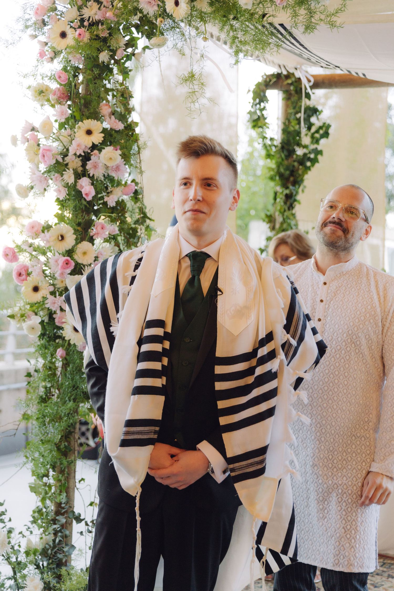 Groom wearing tallit at Jewish wedding ceremony under floral chuppah.