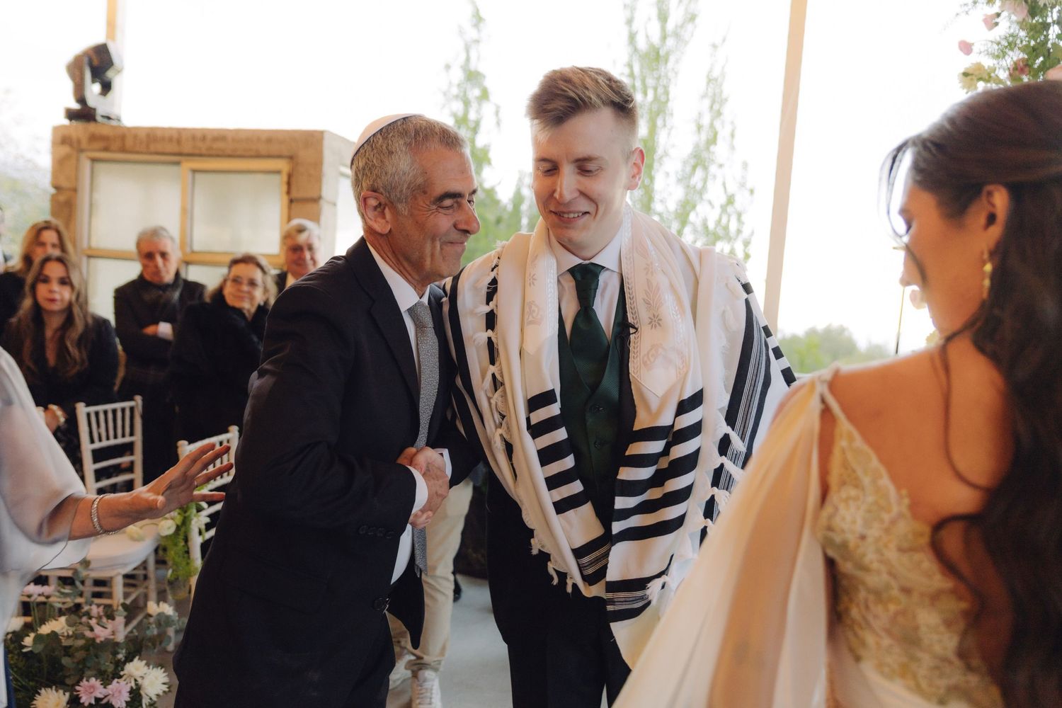 Groom sharing joyful moment with elder under tallit at Jewish wedding.