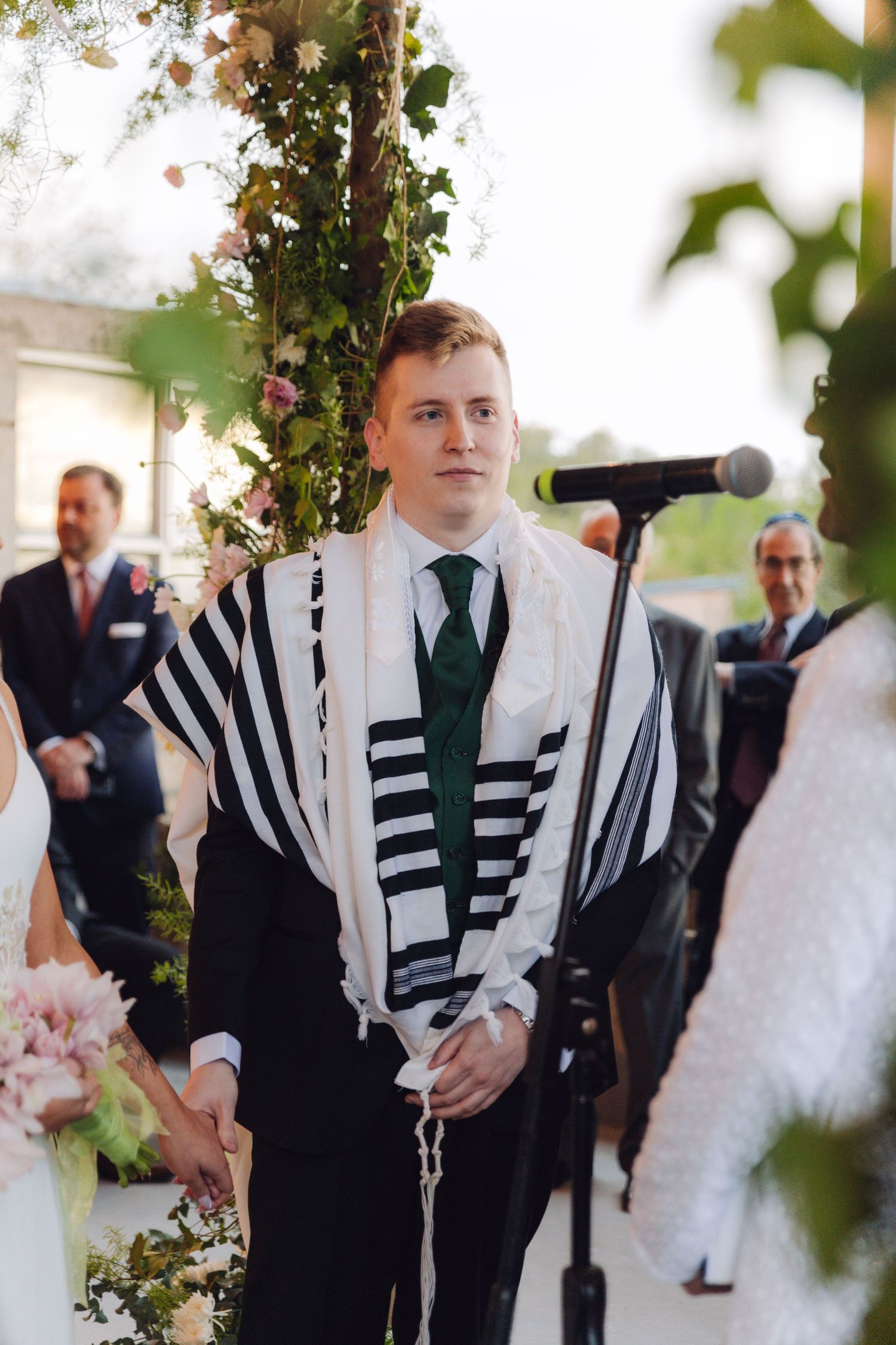 Groom in tallit and green tie stands at floral chuppah awaiting bride.