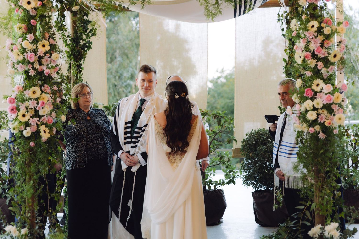 Bride and groom standing together under floral chuppah at Jewish ceremony.
