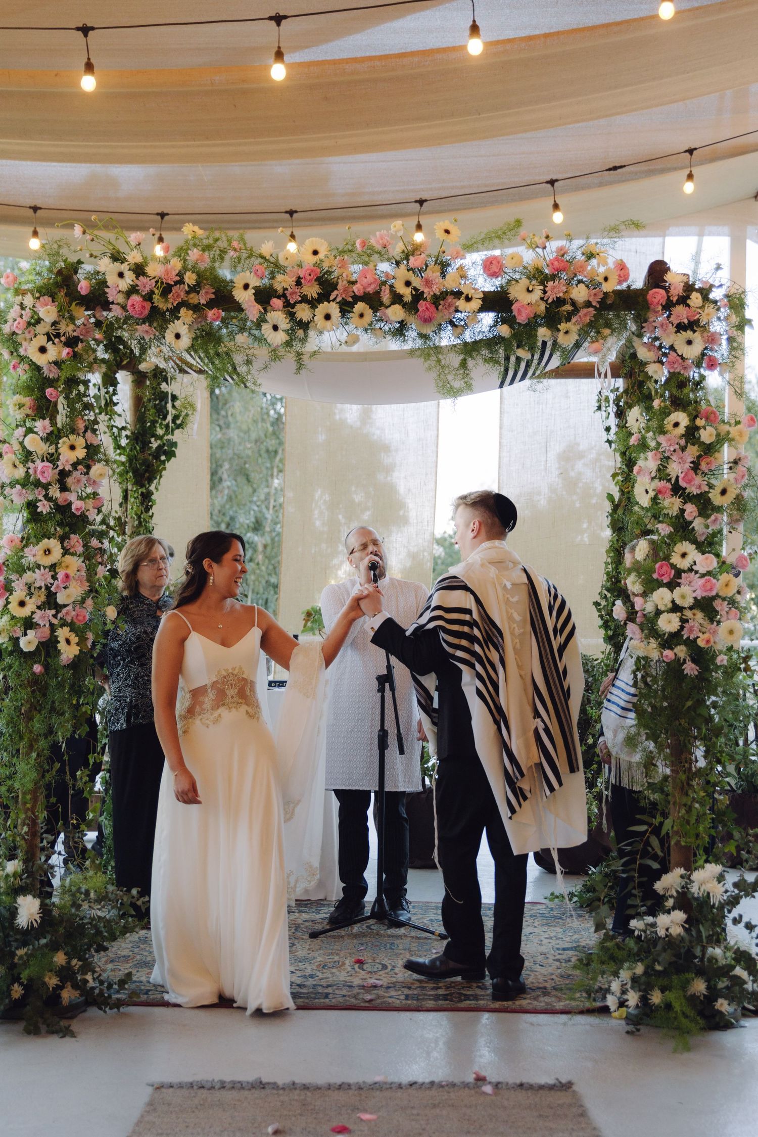 Rabbi officiating Jewish wedding ceremony under pink floral chuppah arch.
