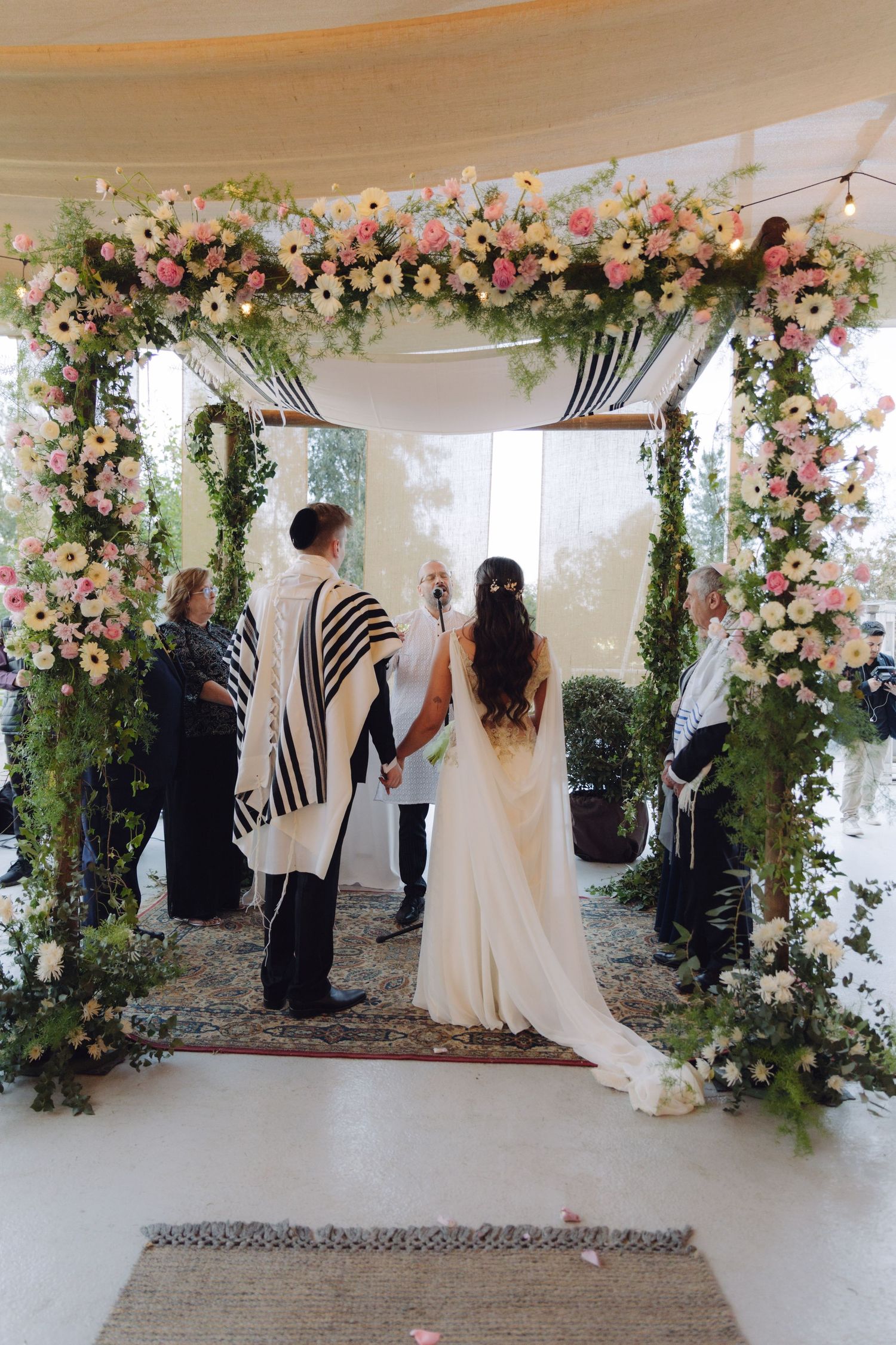 Bride and groom holding hands under lush floral chuppah at wedding.