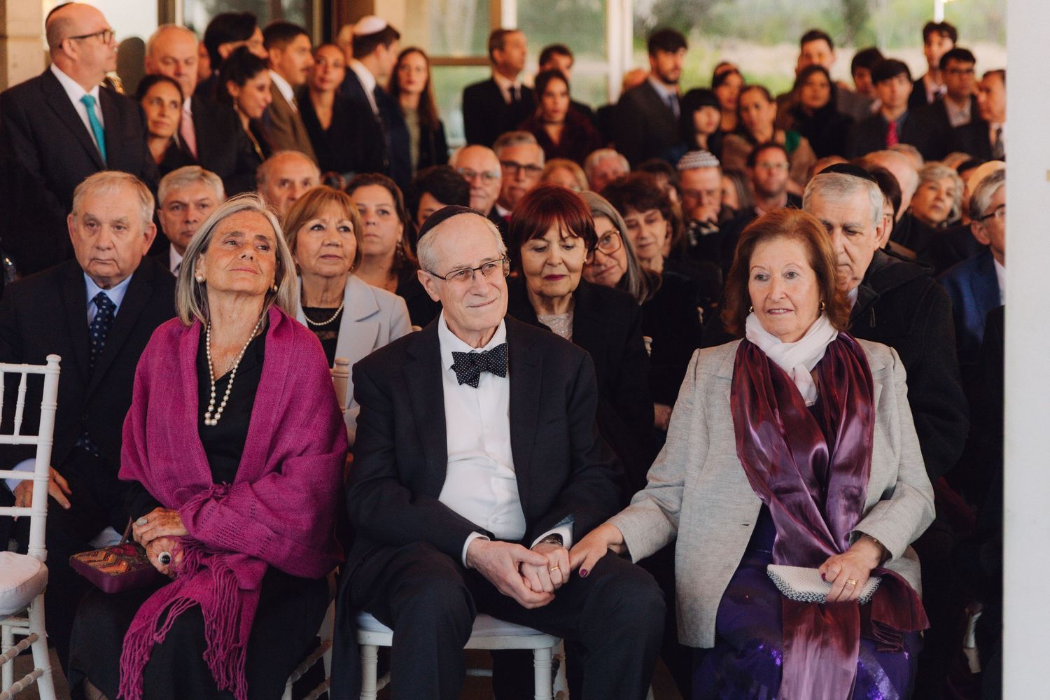 Elegant wedding guests seated and watching Jewish ceremony indoors.