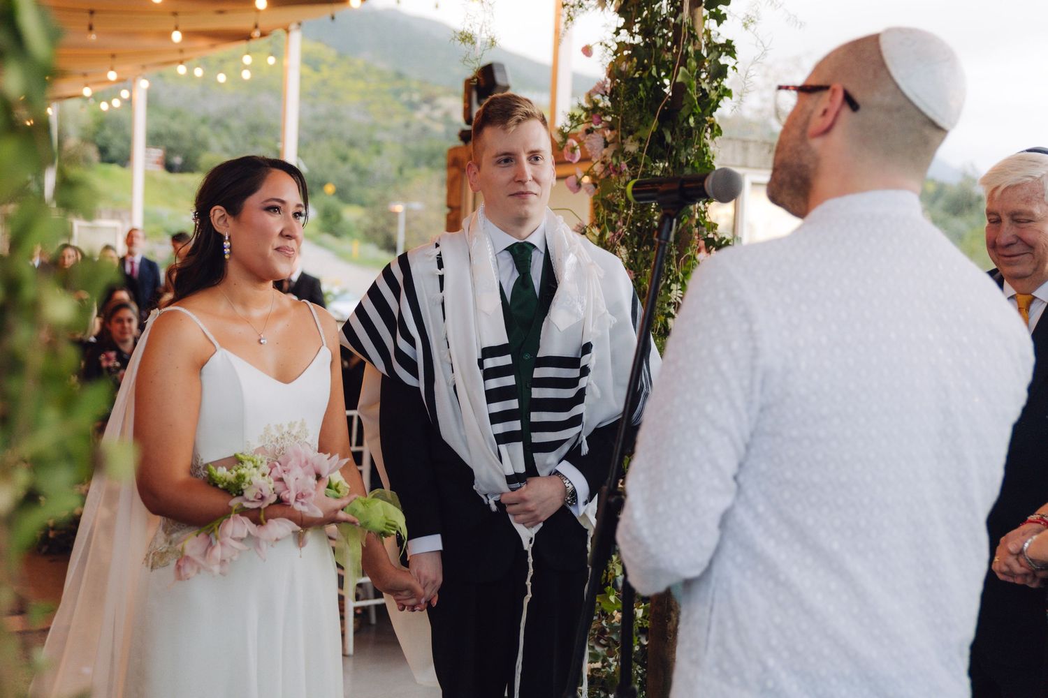 Smiling bride holding bouquet with groom in tallit at Jewish wedding.